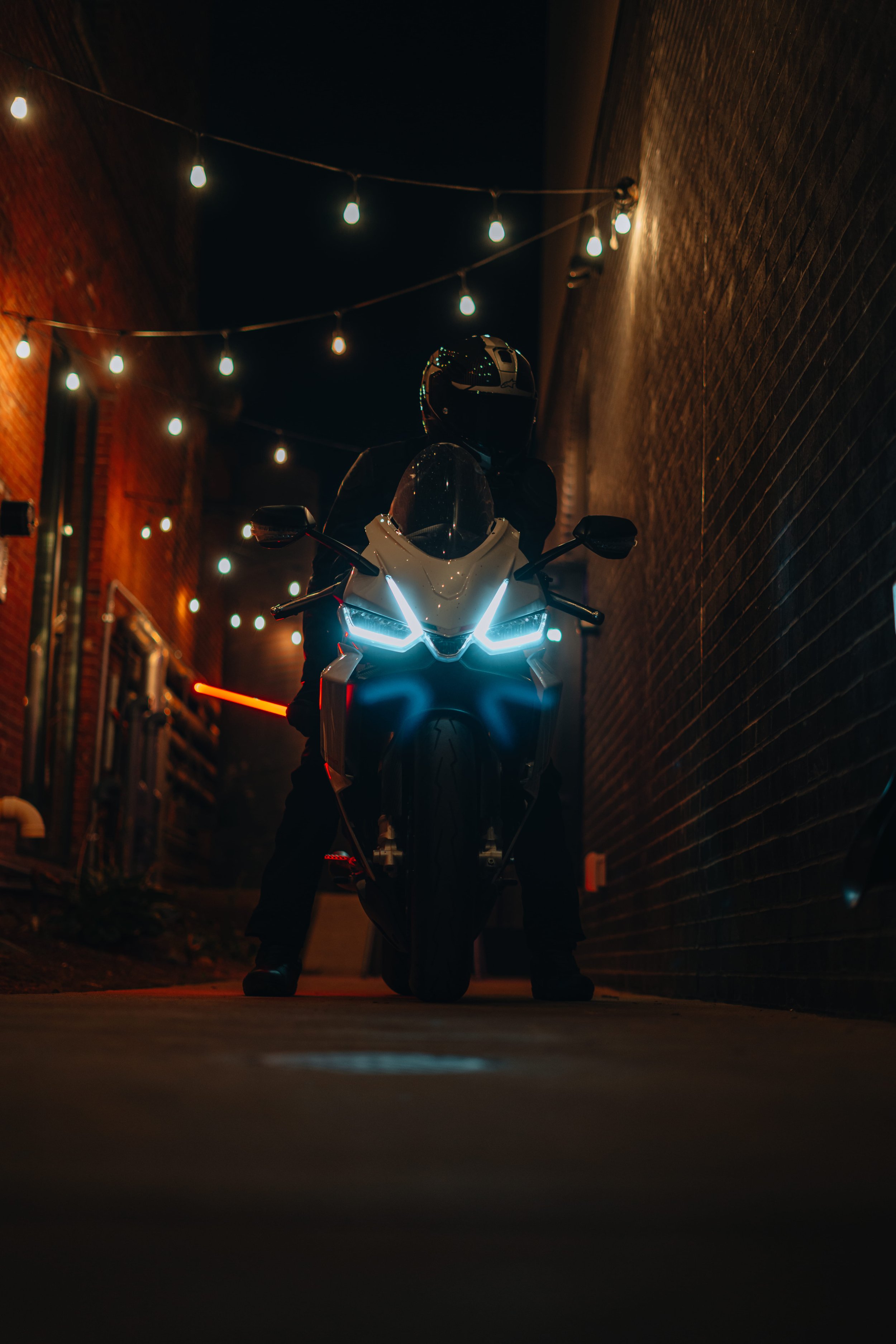 Motorcycle rider wearing helmet at night under string lights, seen from a low angle in an alleyway with brick walls.