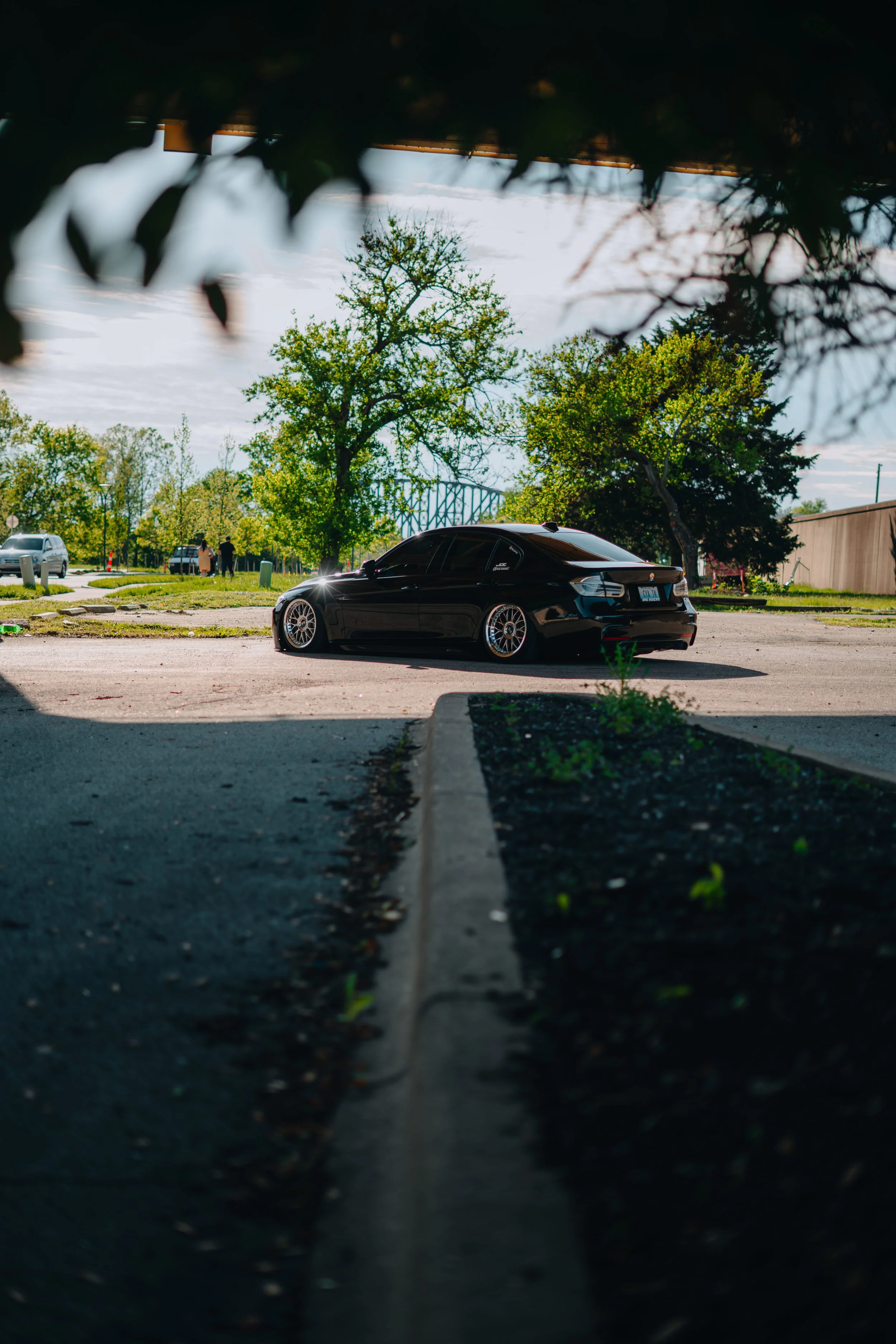 Black car parked in a lot with trees and sky in the background, taken from a low angle through a gap in foliage.
