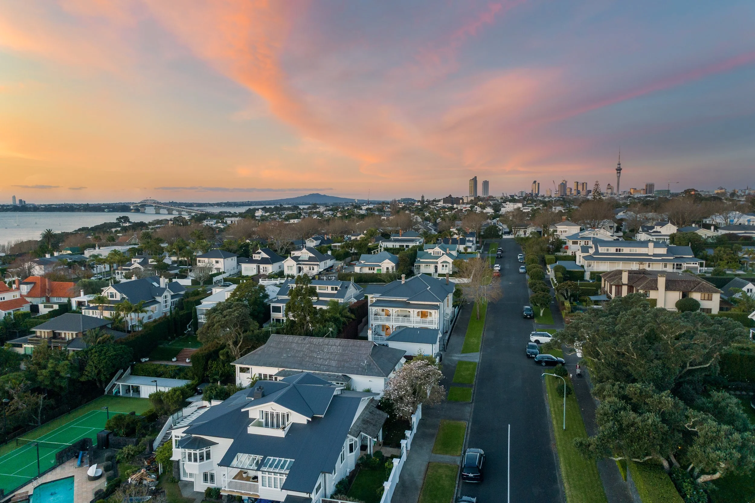 An aerial view of a residential neighborhood near a body of water with a distant city skyline and a tower during pink and orange sunset sky.