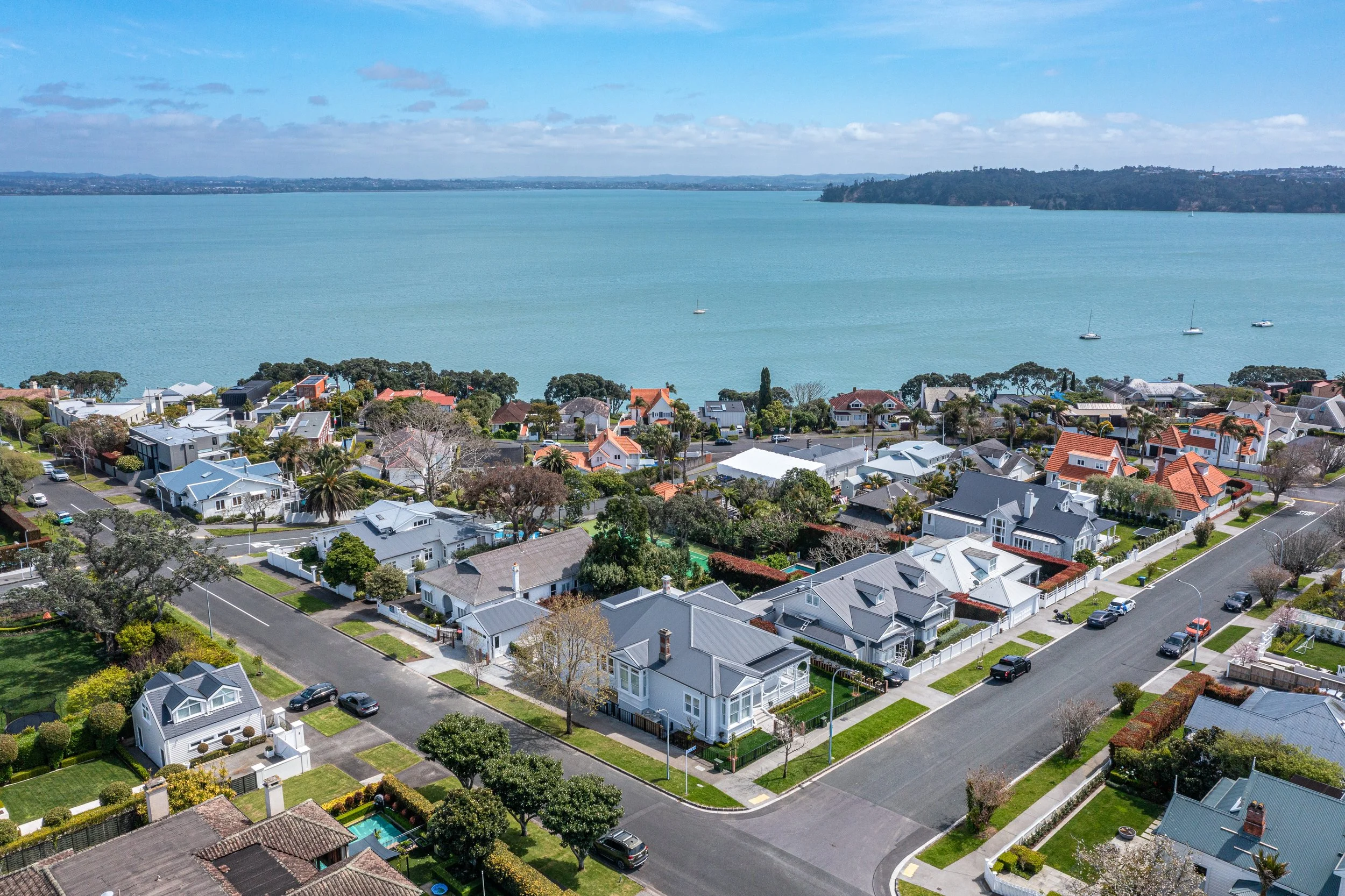 Aerial view of a residential neighborhood by a large body of water, with houses, trees, and streets in the foreground and hills in the background.