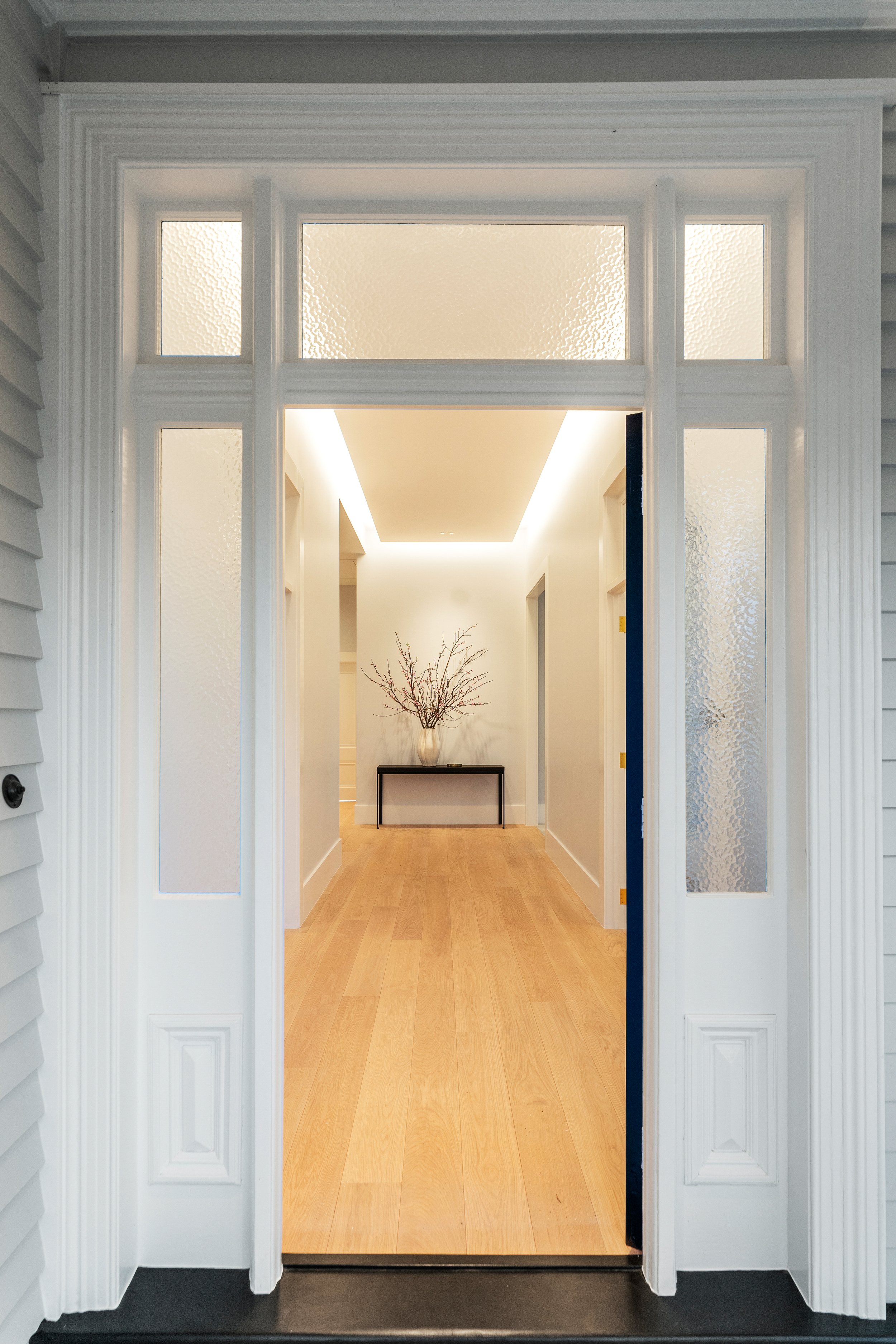 A view through a white front door with glass panels into a brightly lit interior hallway with light wood flooring, white walls, a black console table, and a decorative vase with branches.
