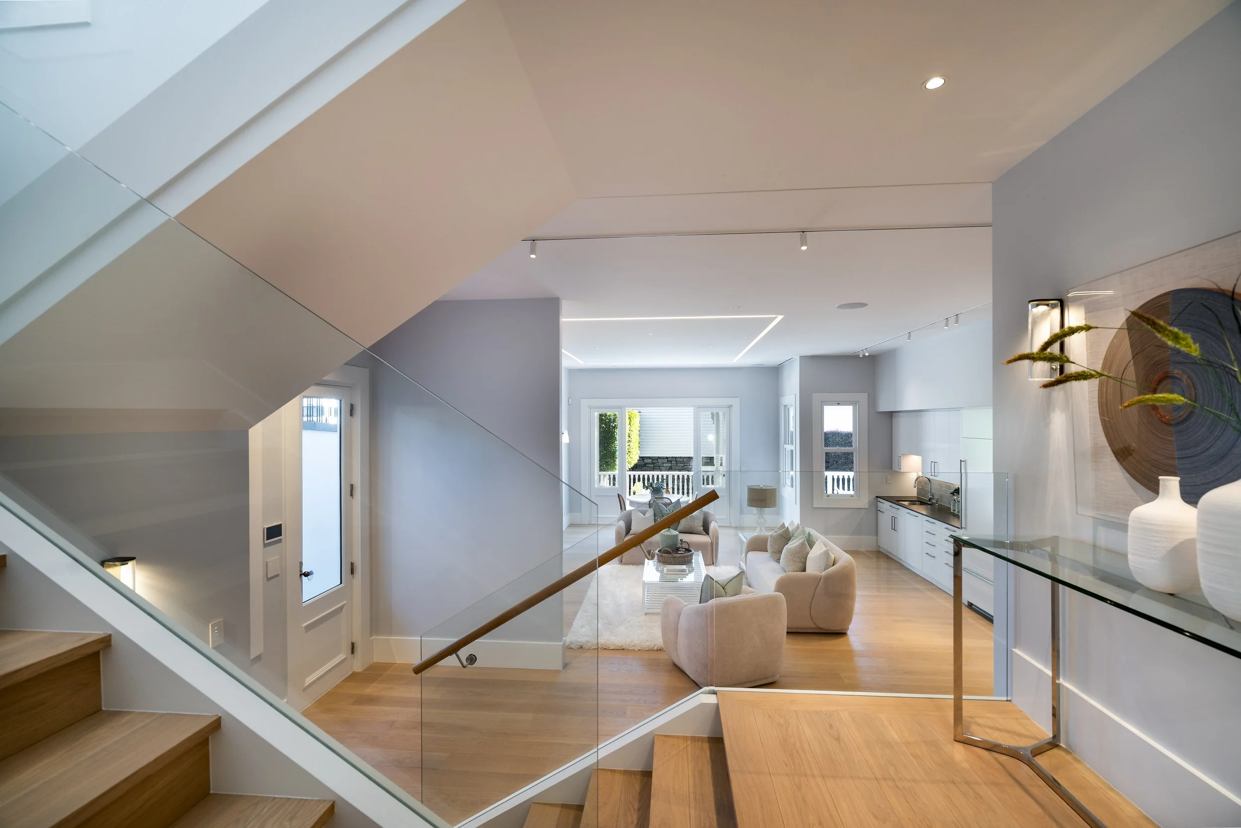 Open-concept living room with staircase and modern decor, including beige sofas, a glass coffee table, artwork, and large windows letting in natural light.