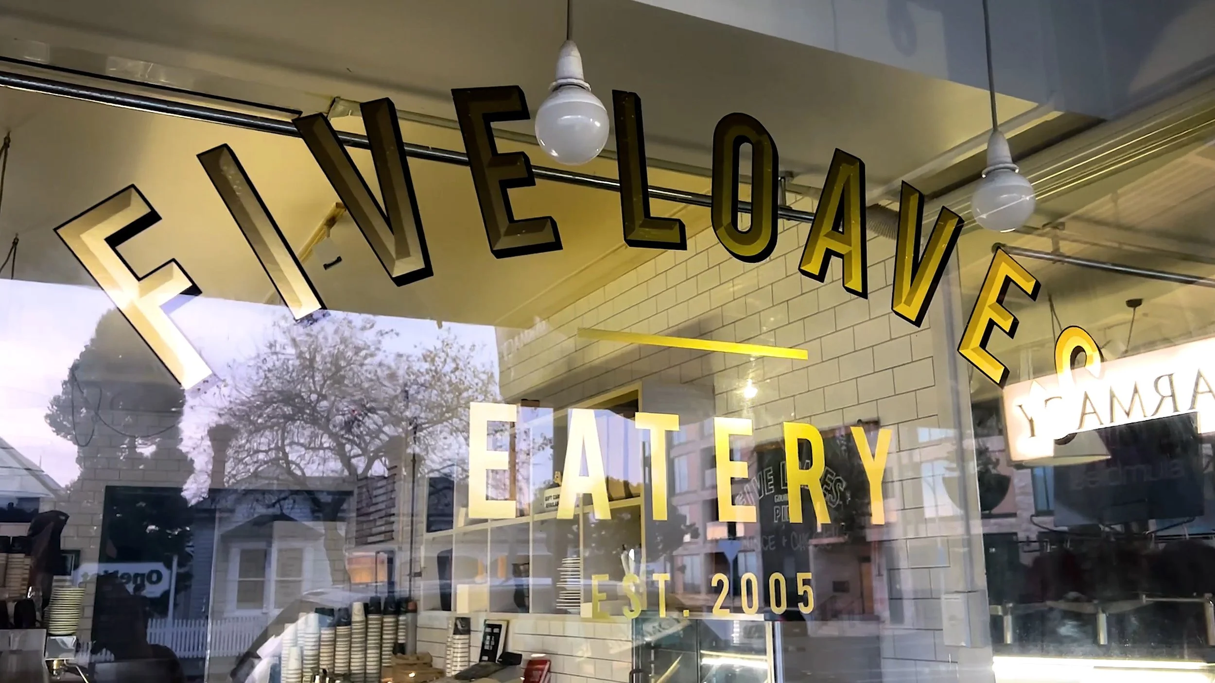 Store window with the words "ELEVEE LOAVES BAKERY" displayed in large black letters with yellow background, reflecting trees and buildings outside.