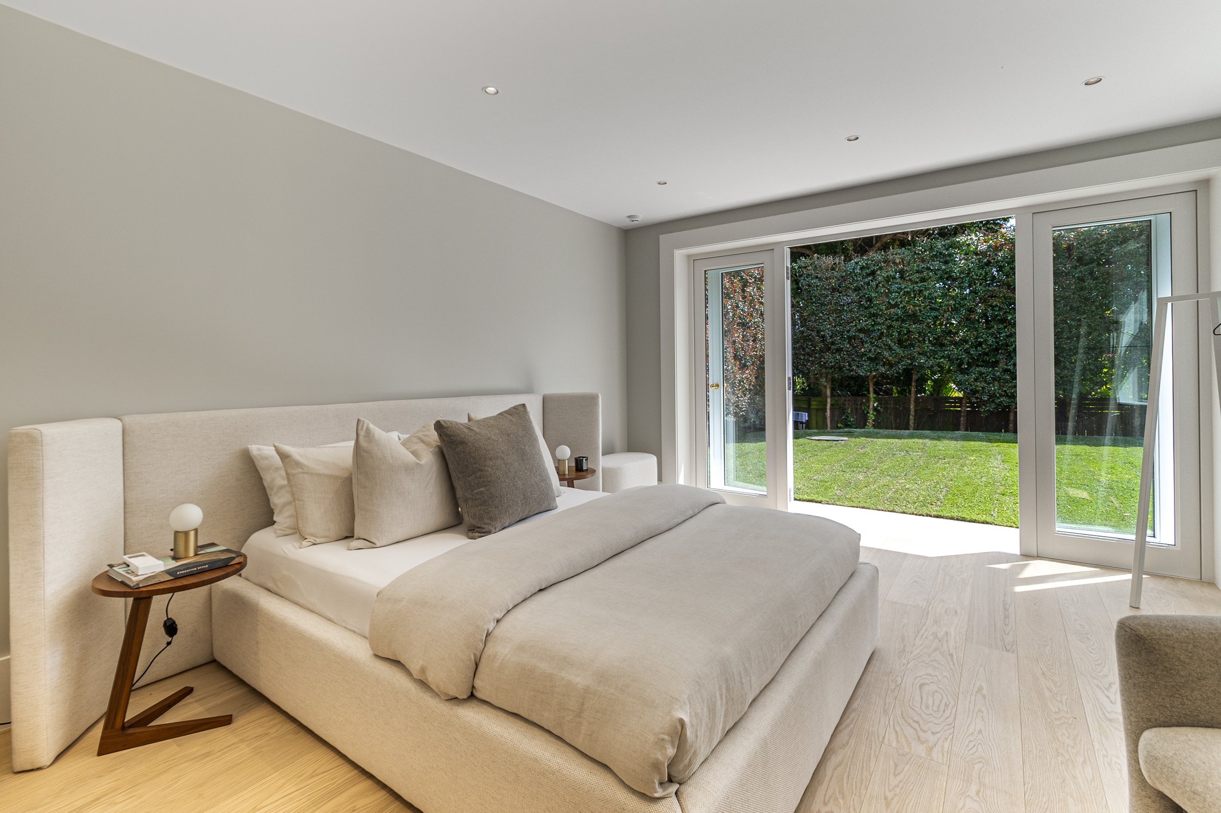 A modern bedroom with a large, light-colored bed, multiple pillows, a side table with books and a lamp, and large glass doors opening to a lush green yard with trees.