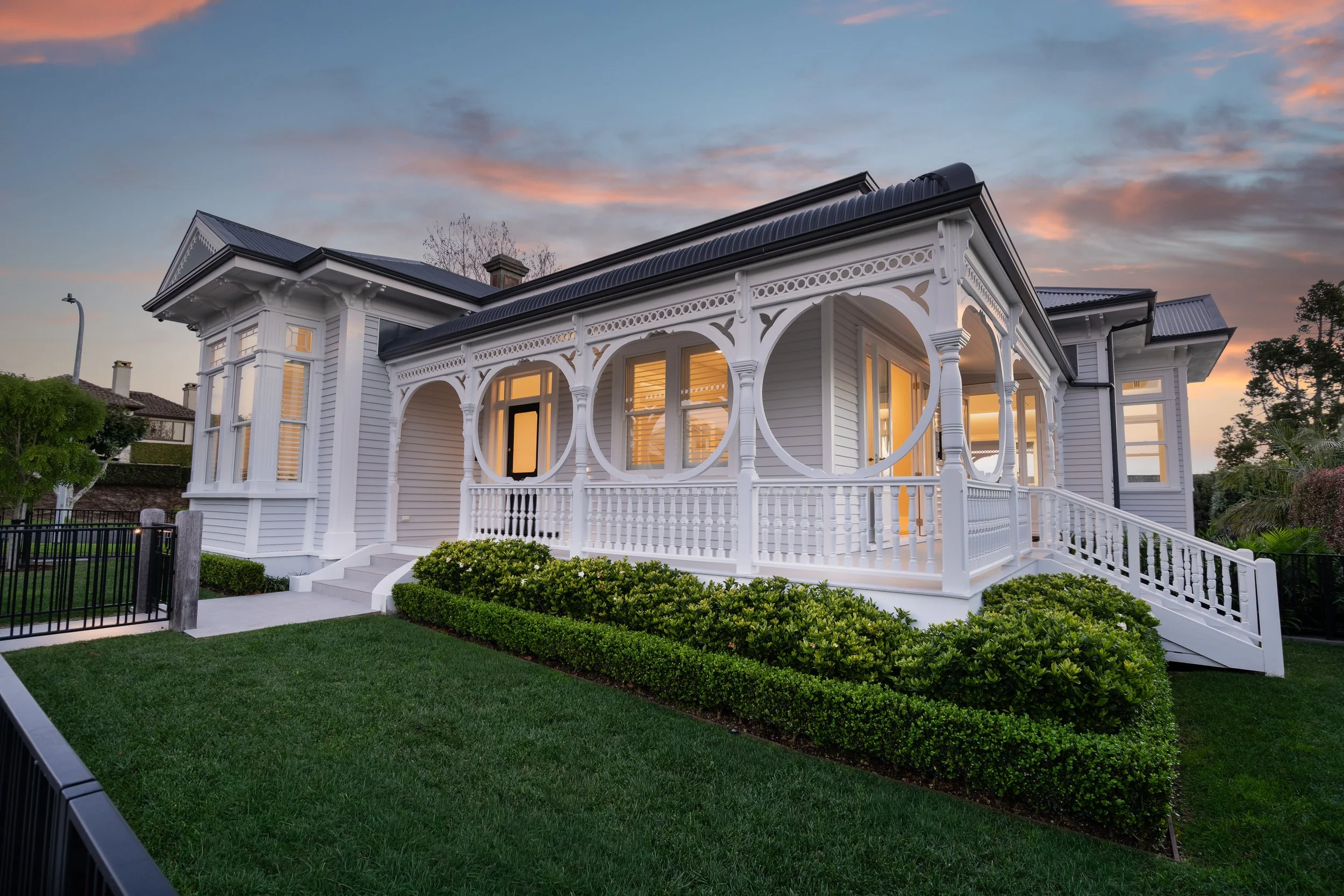 A white house with decorative trim, large windows, and a porch, illuminated from the inside, at sunset. The front yard has trimmed bushes and green grass, with a black fence along the sidewalk. The sky is partly cloudy with a pink and blue hue.