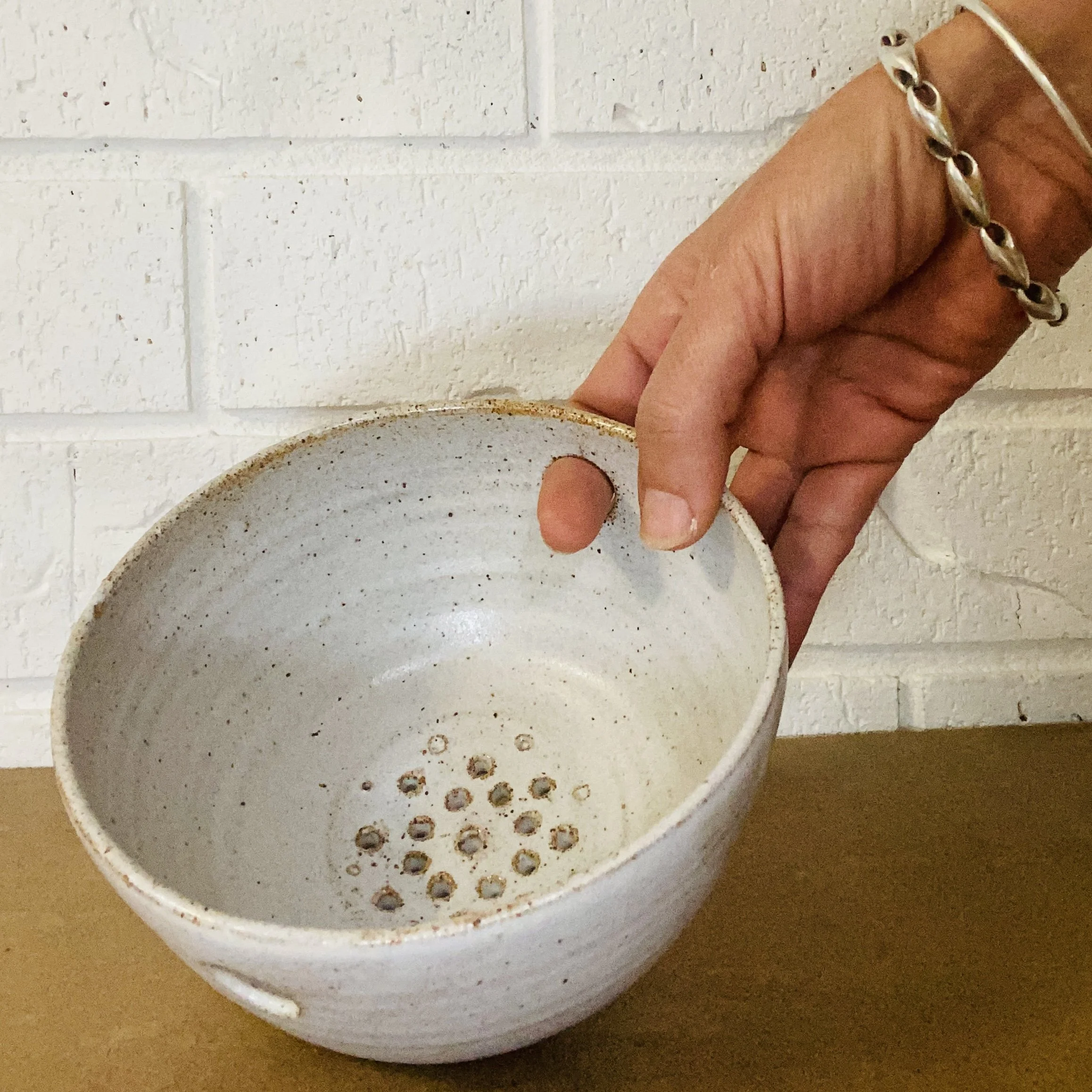 Close-up of a person's hand holding a ceramic or stoneware cup with small holes on the bottom, against a white brick wall background.