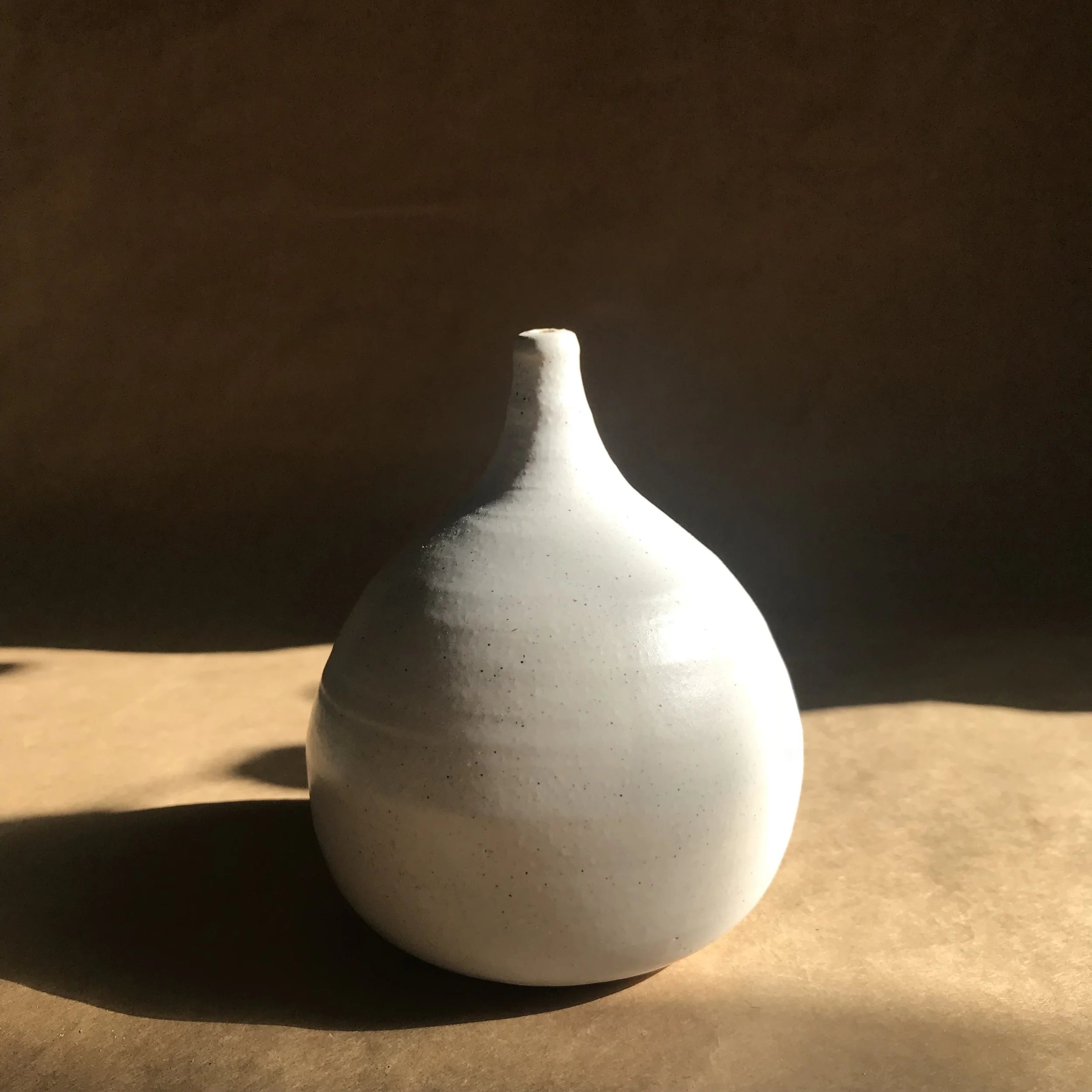 A white ceramic vase with a narrow neck and wider base sitting on a wooden surface, with a dark background.