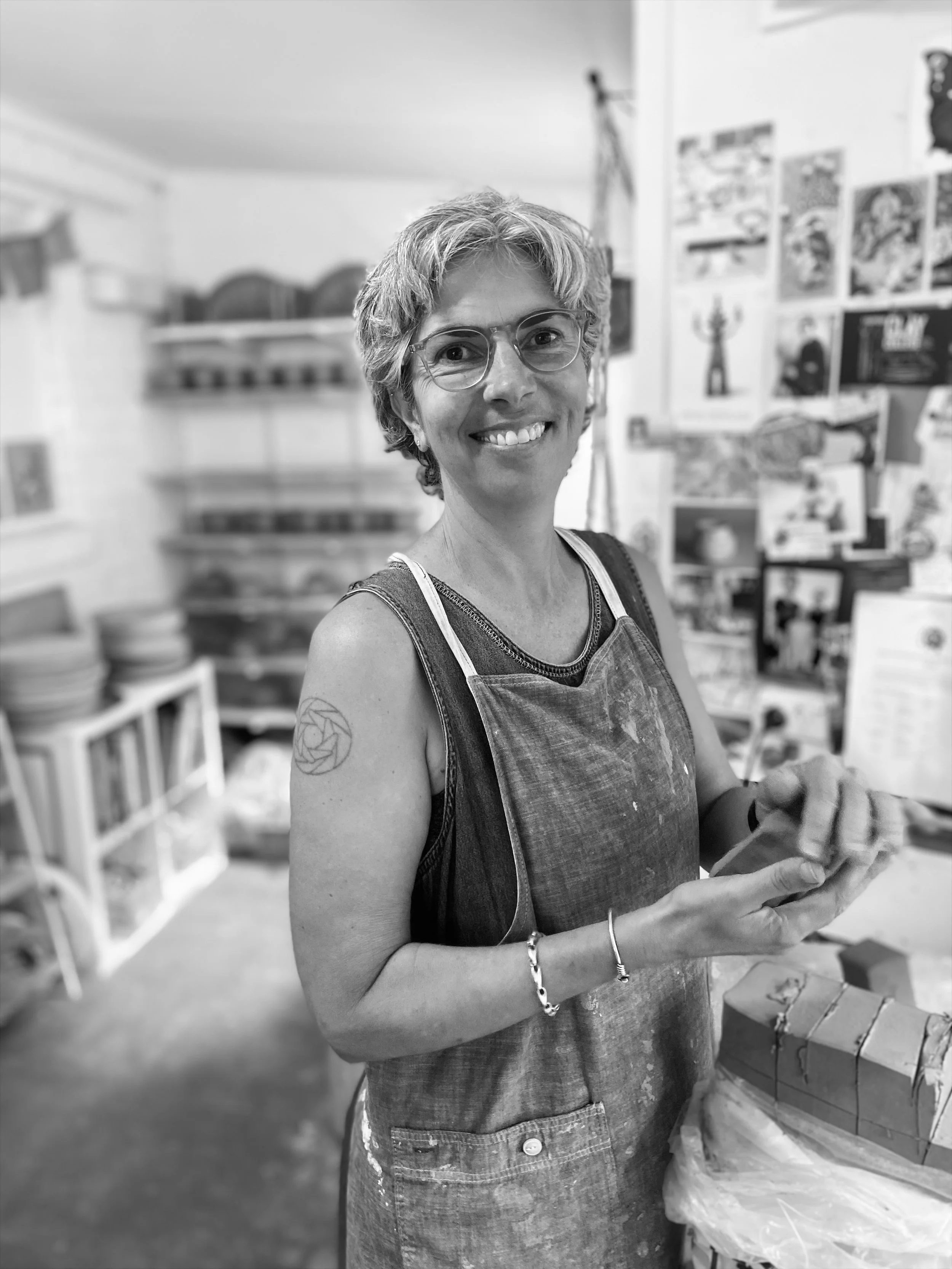 A woman with short, curly hair and glasses smiling in an art or craft studio. She is wearing a sleeveless top and an apron, holding a small object in her hands. The background shows shelves with art supplies and artwork on the walls.