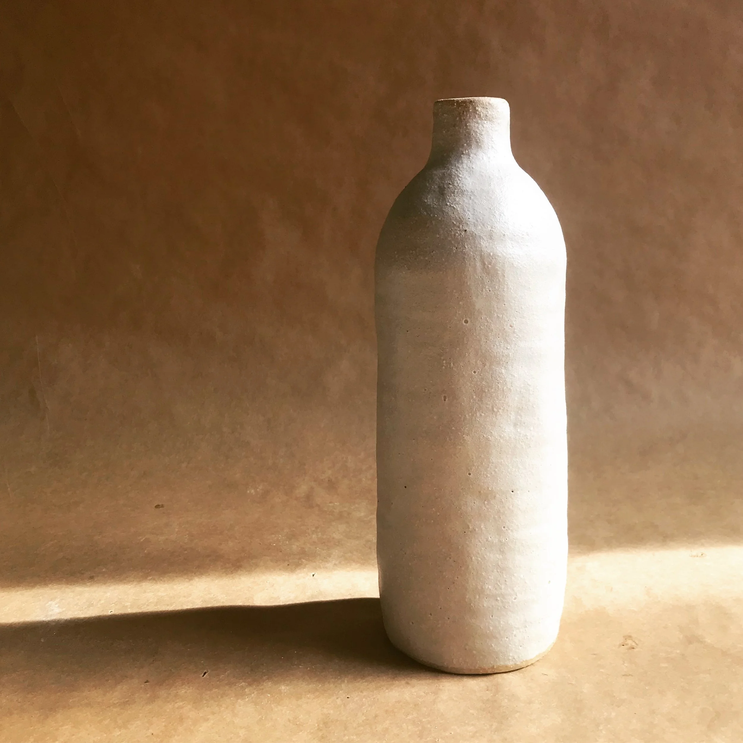 A close-up of a handmade simple, white ceramic vase with a wide body and a narrow neck, situated on a light brown surface against a darker brown background.