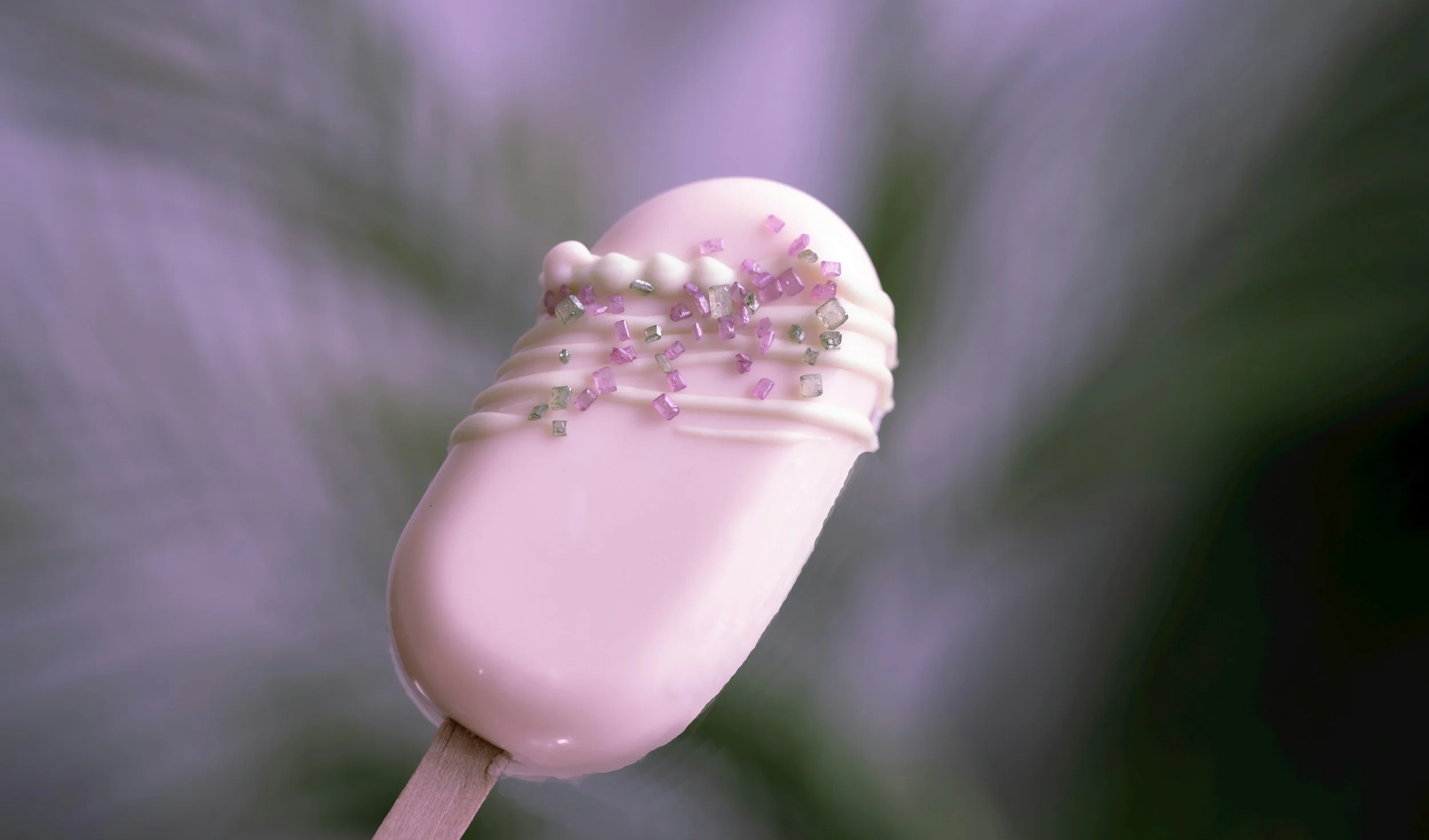Close-up of a light pink ice cream Cakesicles on a wooden stick, decorated with small pink and silver sugar crystals, with a blurred purple and green background.