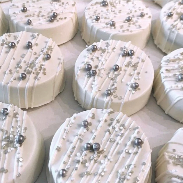 Close-up of round white chocolate dipped Oreos decorated with silver and white sprinkles, metallic silver spherical decorations, and white chocolate drizzle.