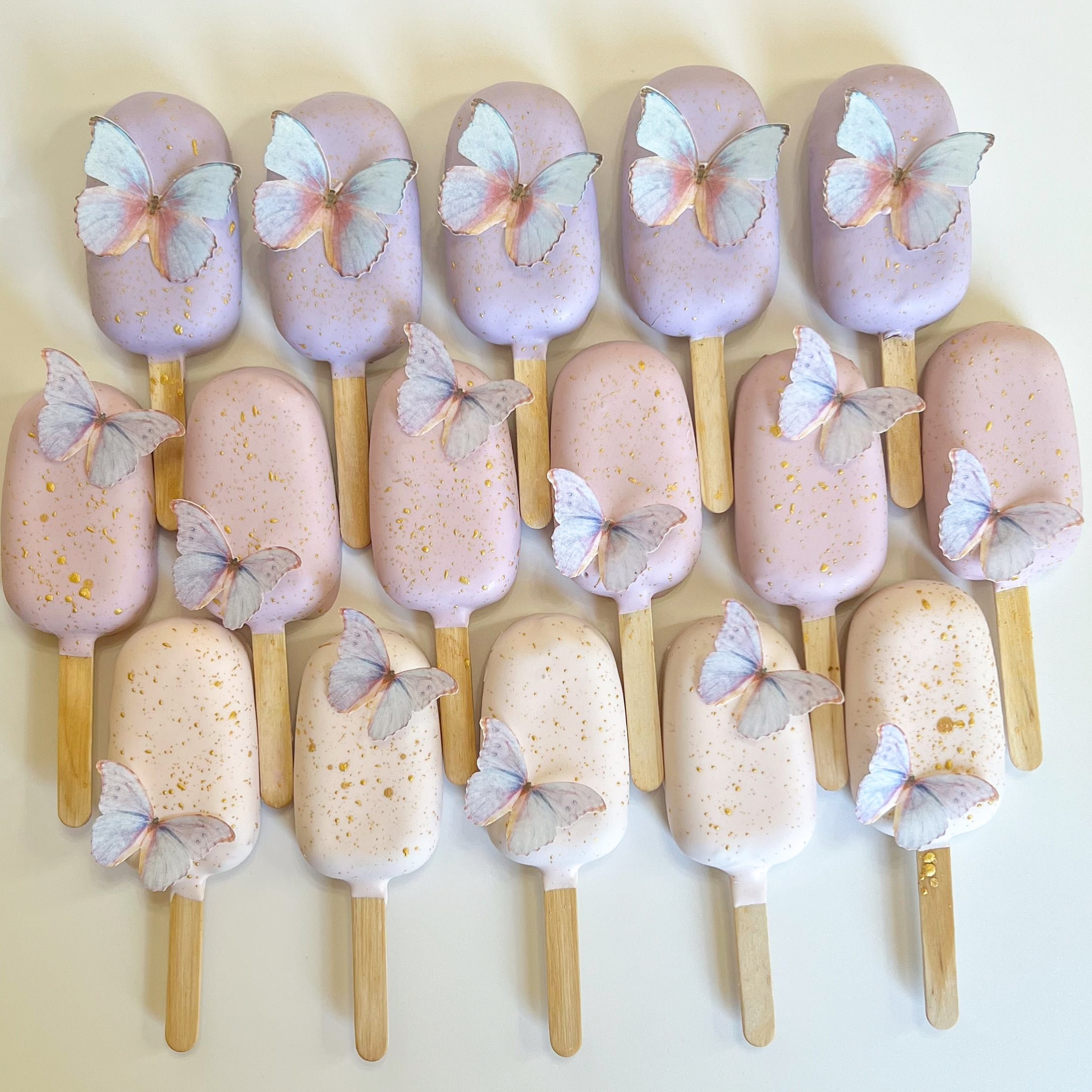 Assorted pastel cakesicles decorated with white and pink butterfly toppers and gold flecks, arranged in two rows on a white background.
