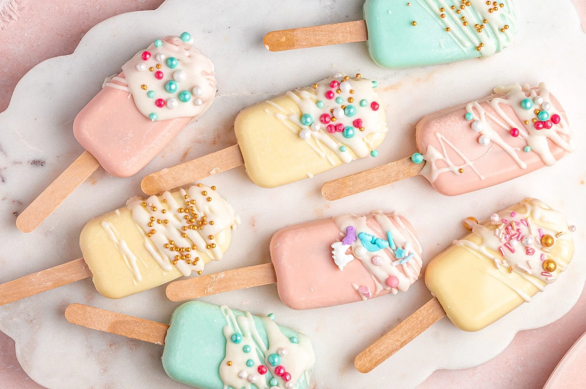 Colorful pastel cakesicles decorated with sprinkles and white icing on a white surface.