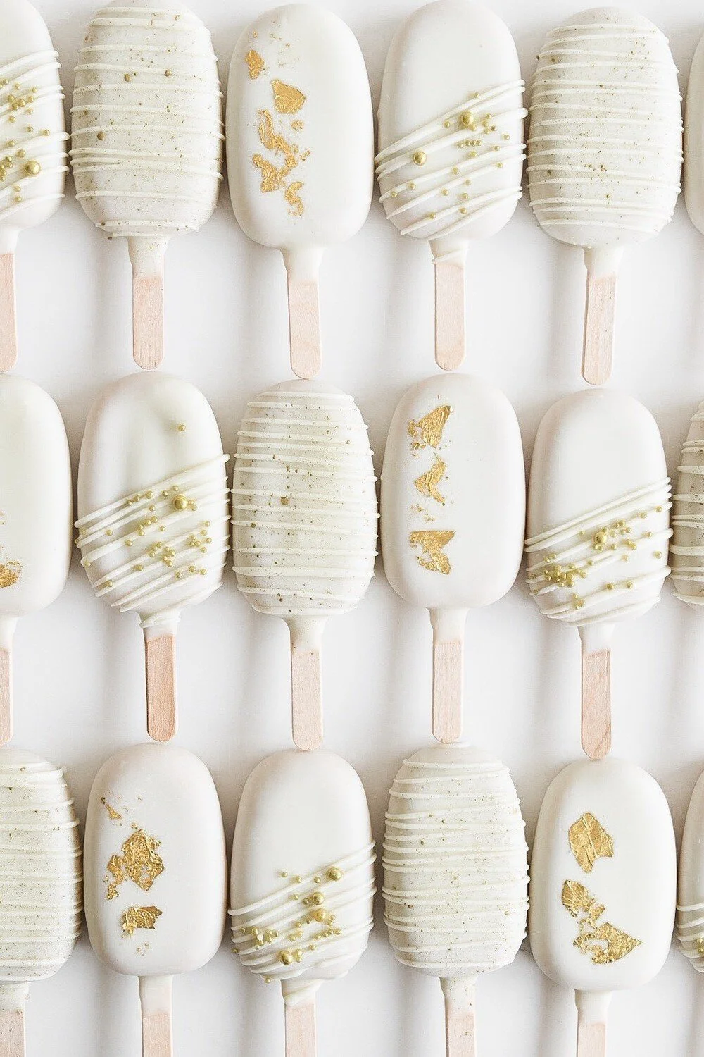 White chocolate-dipped cakesicles decorated with gold leaf and pearl-like edible beads, arranged in neat rows.