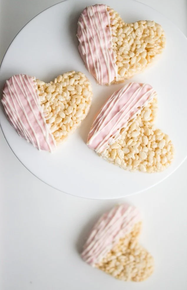Four heart-shaped rice crispy treats with pink and white drizzle on a white plate, one treat is on the table.