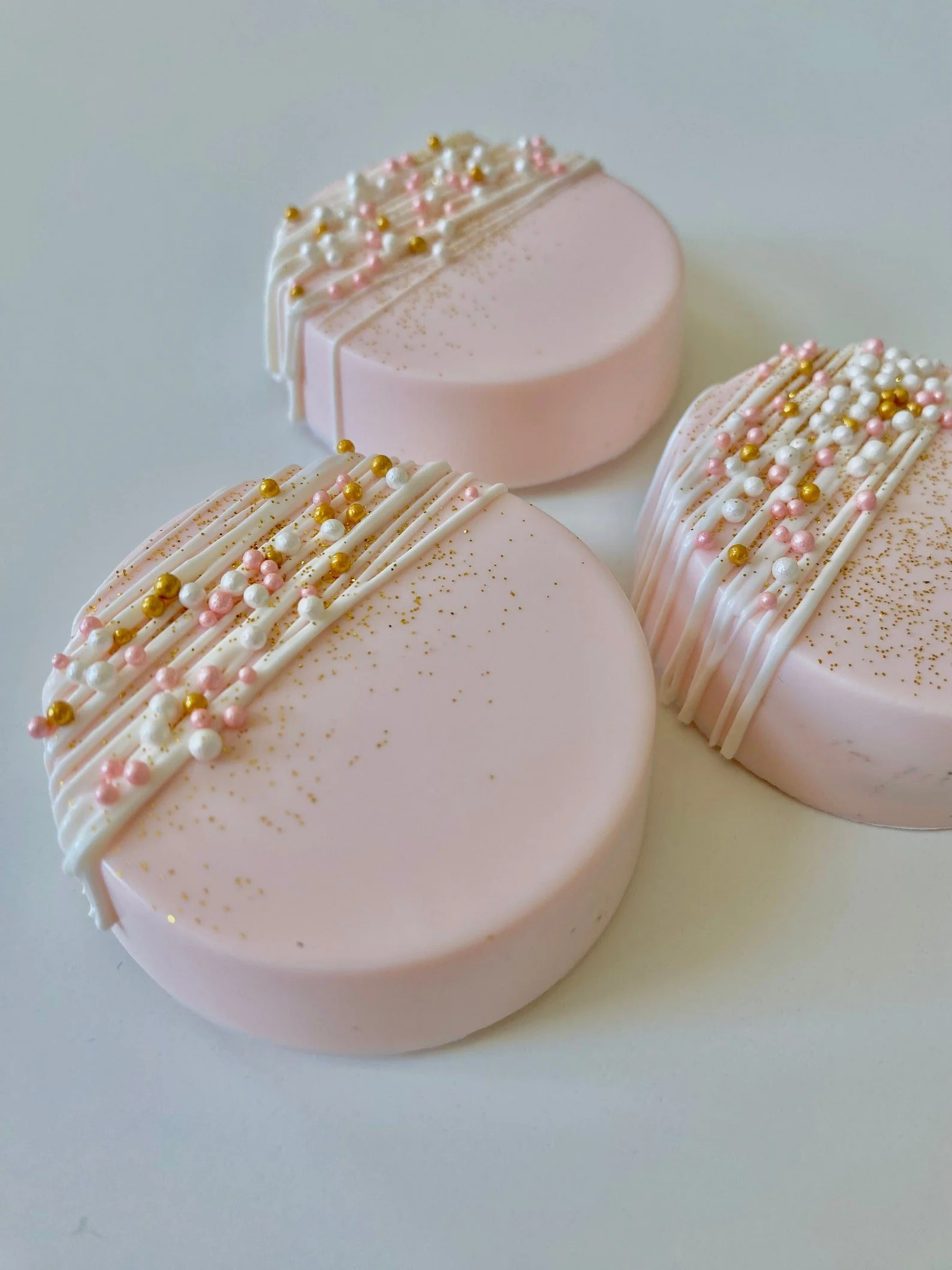 Three pink round chocolate dipped Oreos decorated with white, pink, and gold sprinkles and white drizzle on top.