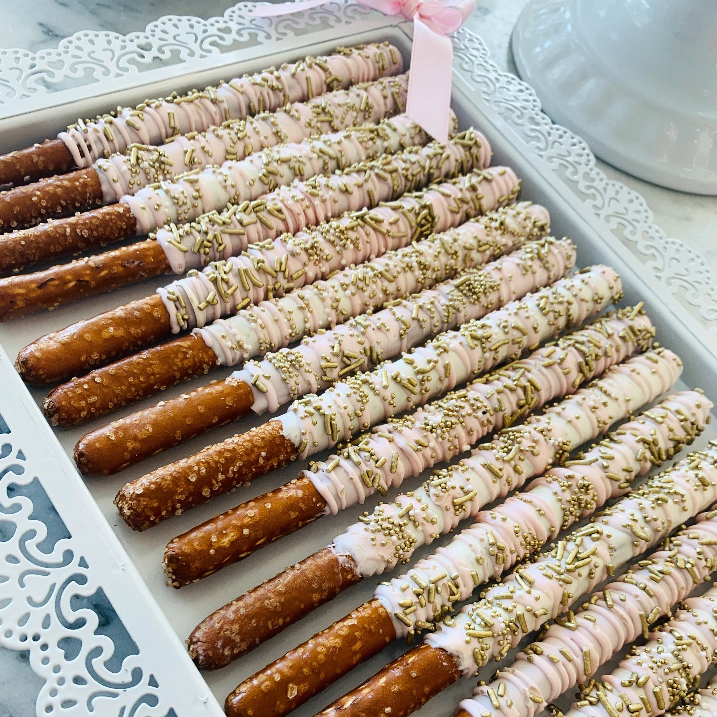 A decorative white tray containing several pretzel rods covered in white pink icing and topped with gold sprinkles.