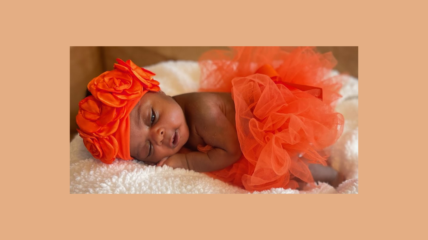 A newborn baby girl lying on a white blanket, wearing an orange tulle dress and matching headwrap with large roses.