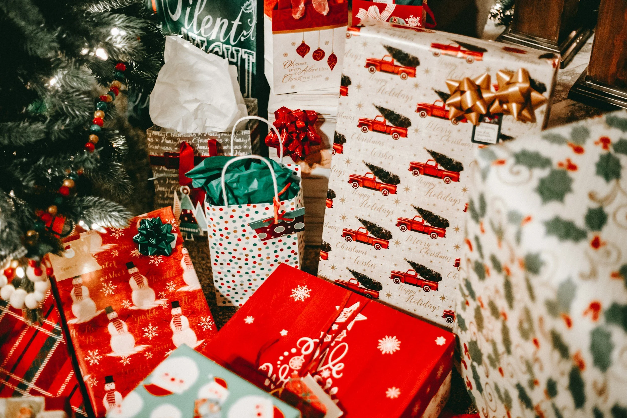 Several wrapped Christmas presents in festive paper with red, white, and green designs under a Christmas tree decorated with lights and ornaments.
