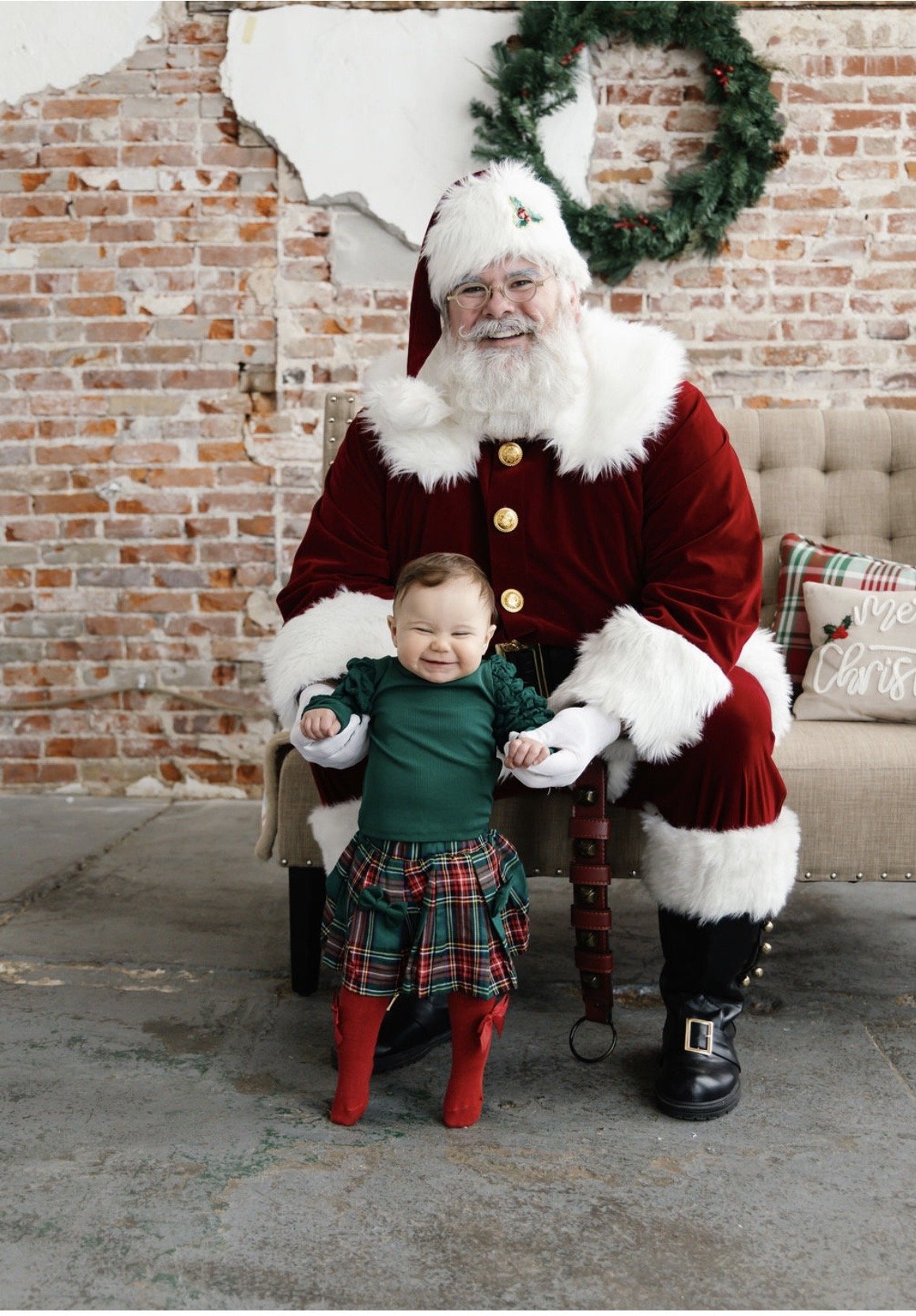 Santa Claus in a red velvet suit with white fur trim, sitting on a beige sofa with a young girl wearing a green top, a plaid tartan skirt, red tights, and red boots. They are indoors with a brick wall and holiday decorations like a wreath and Christm
