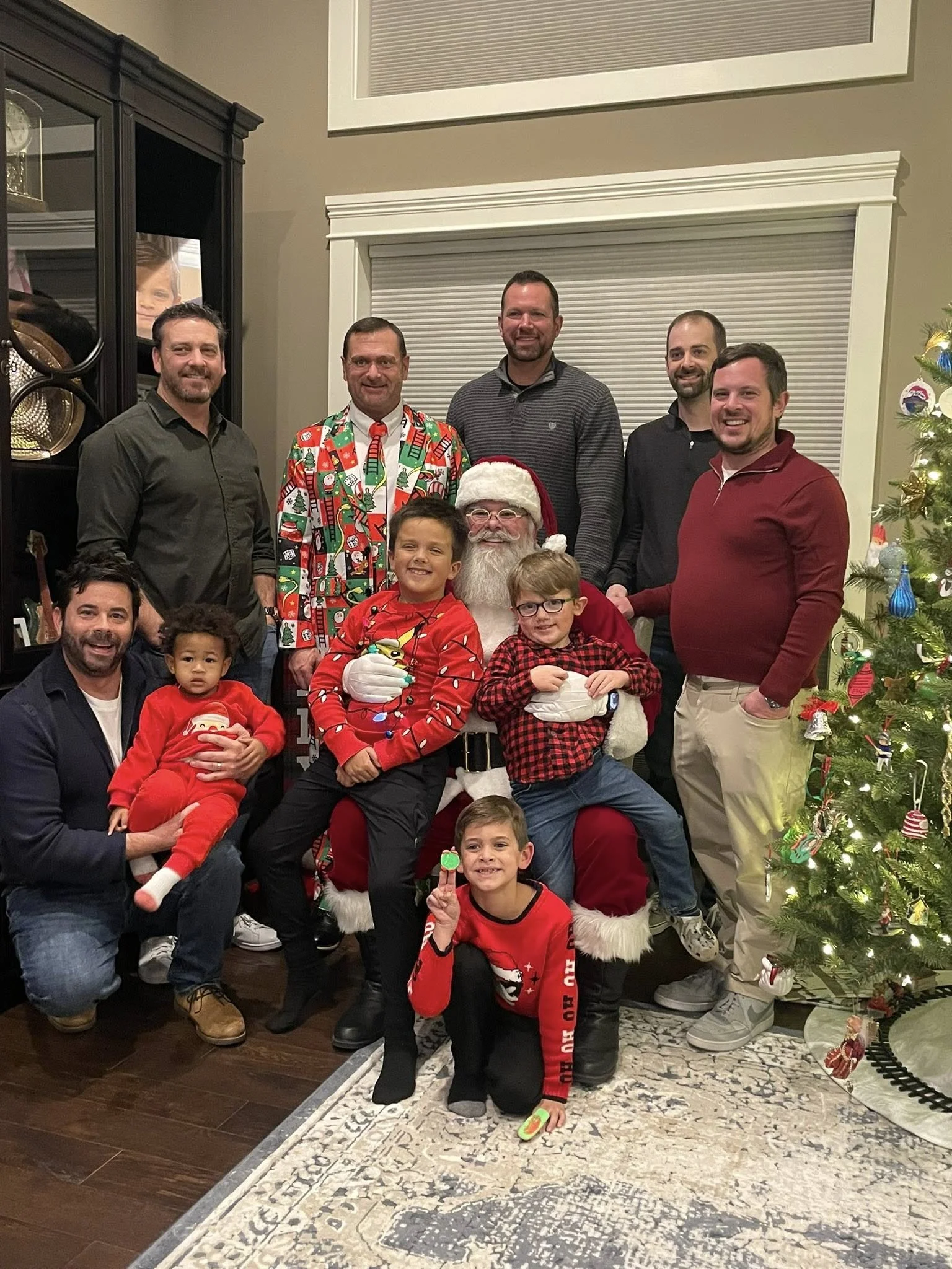 Group photo of children and adults with Santa Claus during a Christmas celebration in a living room, with a decorated Christmas tree on the right.