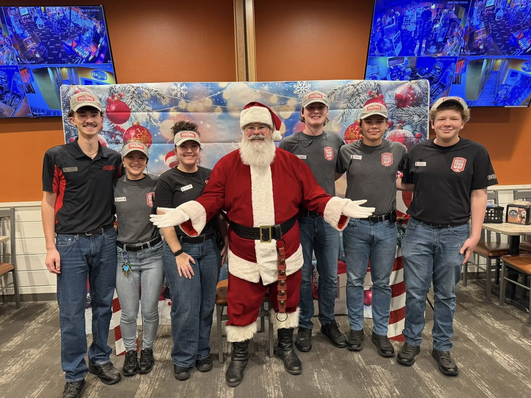 Group of six young people and one man dressed as Santa Claus, standing in front of a holiday-themed backdrop with Christmas ornaments, pine branches, and snowflakes. All are smiling and wearing black shirts and caps with a restaurant logo.