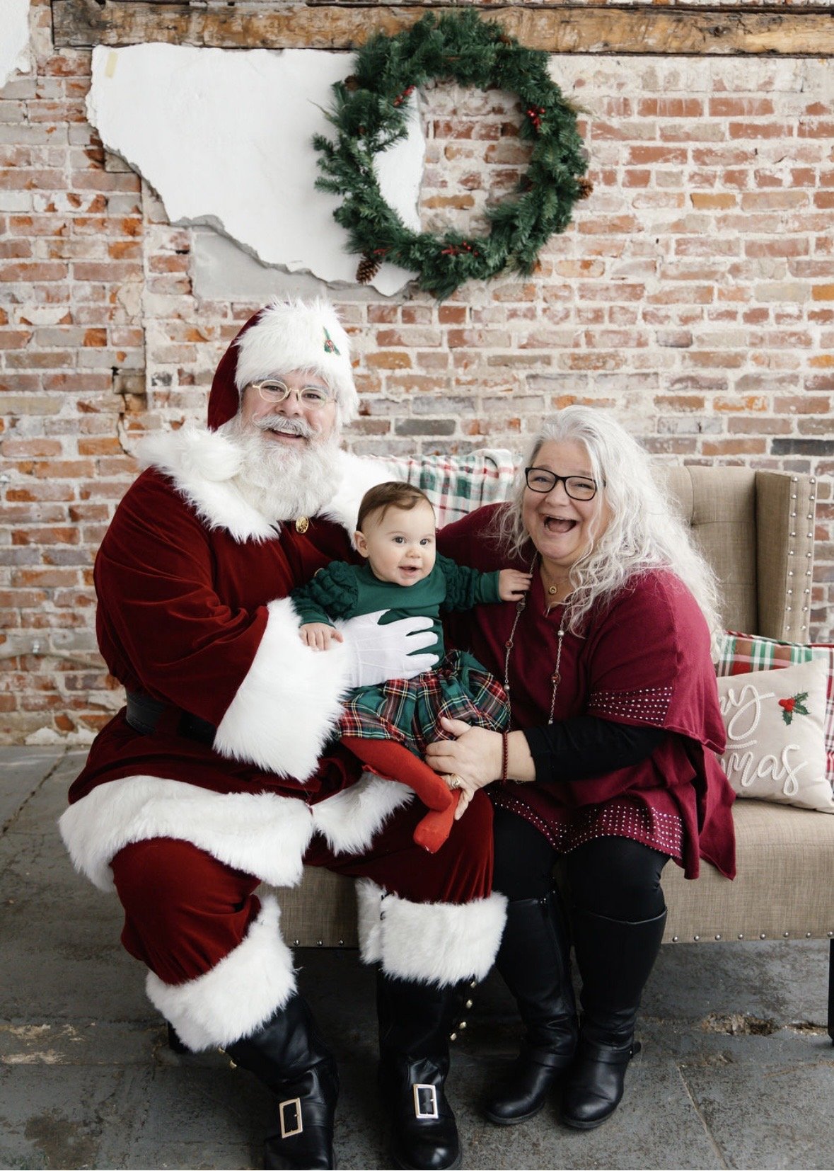 A family celebrating Christmas, featuring a man dressed as Santa Claus holding a baby, and a woman sitting beside them, all smiling happily. The background includes a brick wall with a Christmas wreath and holiday decorations.