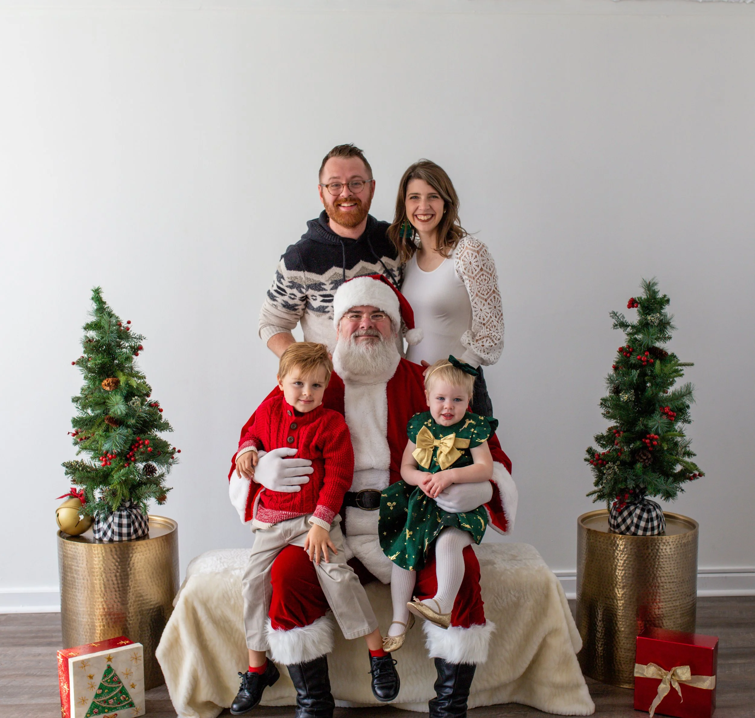 A group of five people, including a man dressed as Santa Claus, two children, and two adults, posing for a holiday photo. They are standing in front of a plain white wall with Christmas trees and presents around them.
