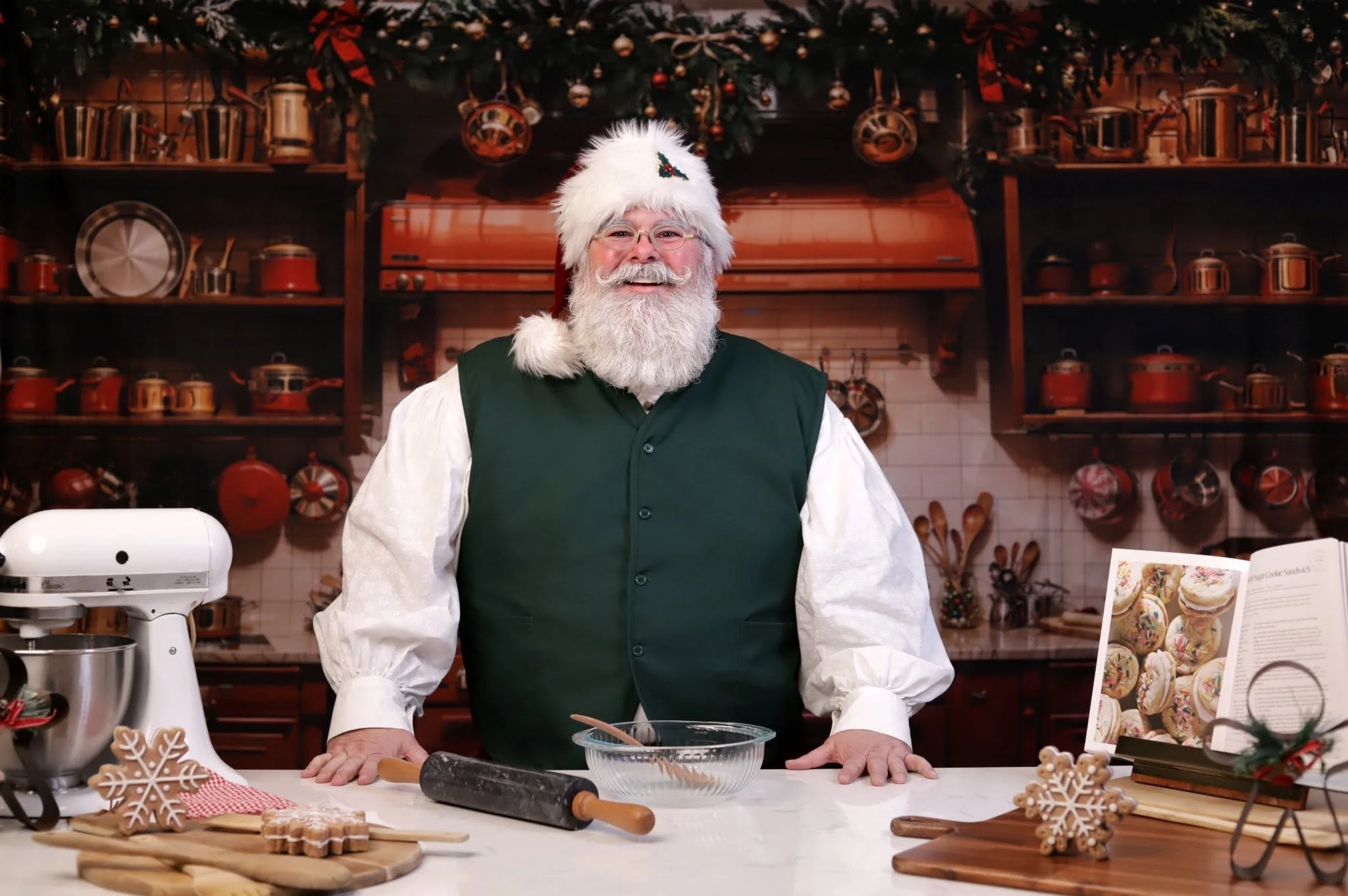 A cheerful man dressed as Santa Claus with glasses, a white beard, and a Santa hat, standing in a kitchen decorated for Christmas. The kitchen features wooden shelves with pots and decorations, and holiday cookies on the counter.