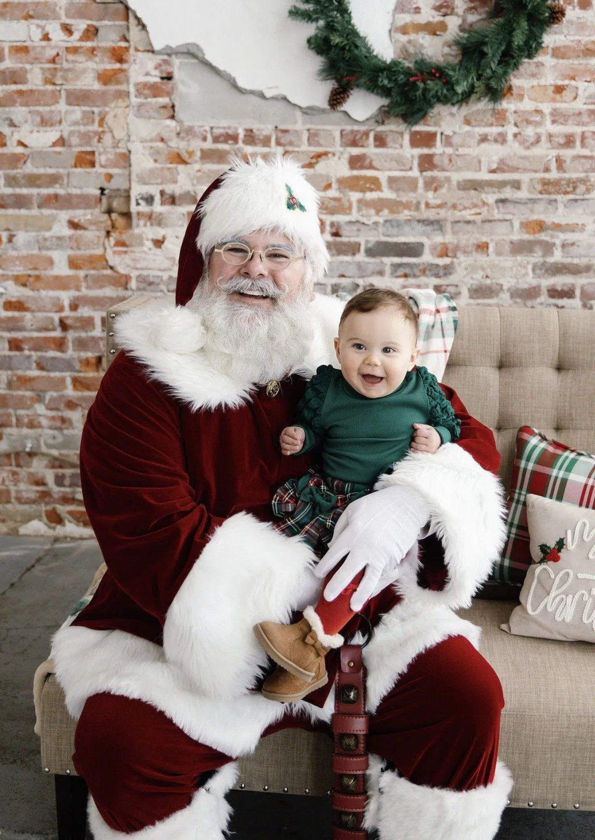 Santa Claus with glasses and a beard, dressed in a red velvet suit with white fur trim, sitting on a beige chair, holding a smiling baby in a green top and plaid pants, in a festive holiday setting with a brick wall and Christmas wreath in the backgr