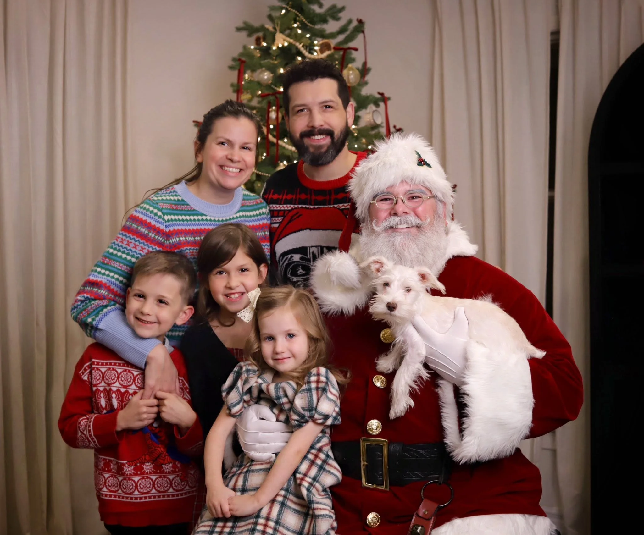 Family portrait with children, seated with Santa Claus and a woman, in front of a Christmas tree decorated with lights, ornaments, and red ribbons.
