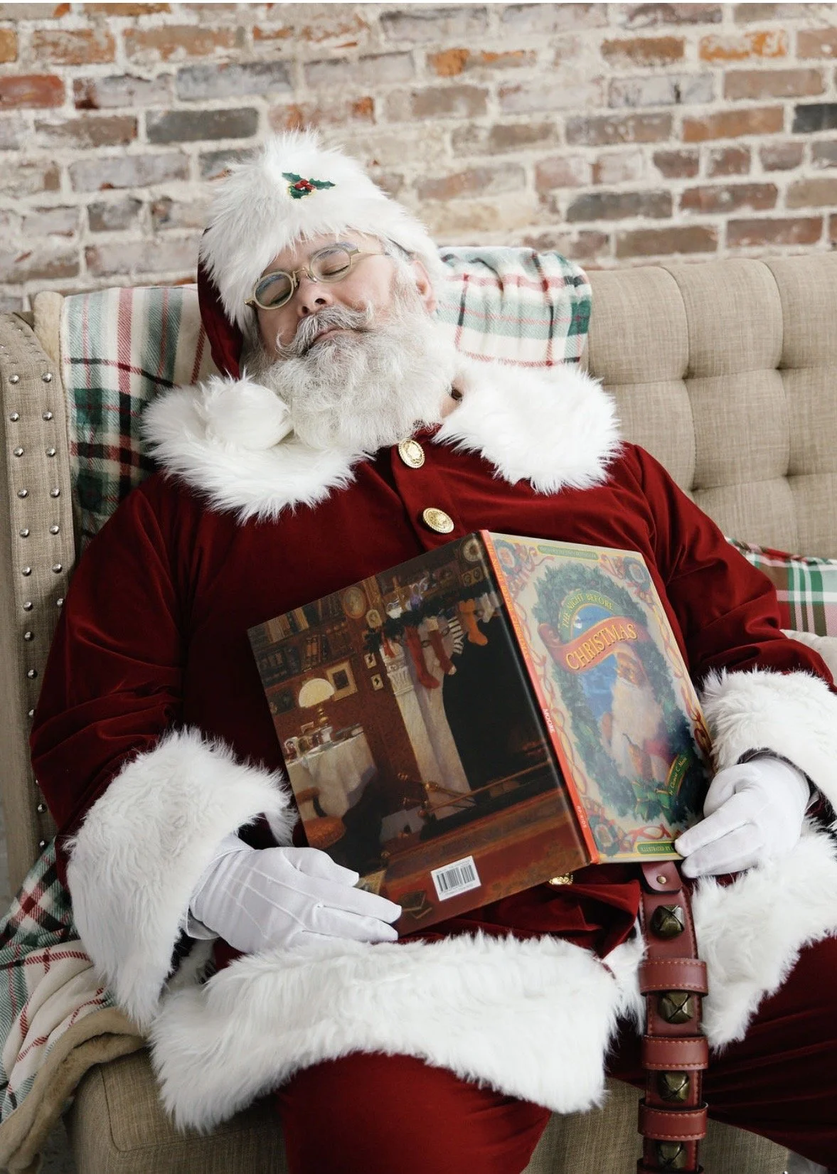 Santa Claus wearing a red velvet suit with white fur trim, a Santa hat with a holly decoration, and glasses, is peacefully sleeping with a Christmas-themed book in his lap, sitting on a beige tufted sofa.
