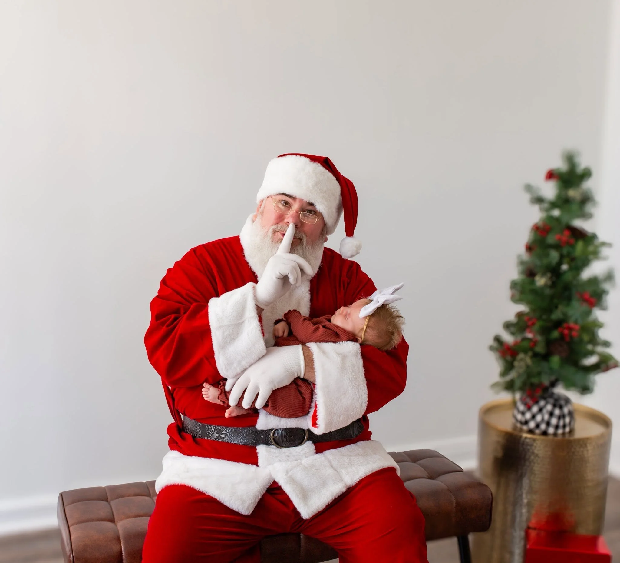Santa Claus with glasses and a Santa hat, holding a finger to his lips, standing in a room with a small decorated Christmas tree and a plant in a planter.