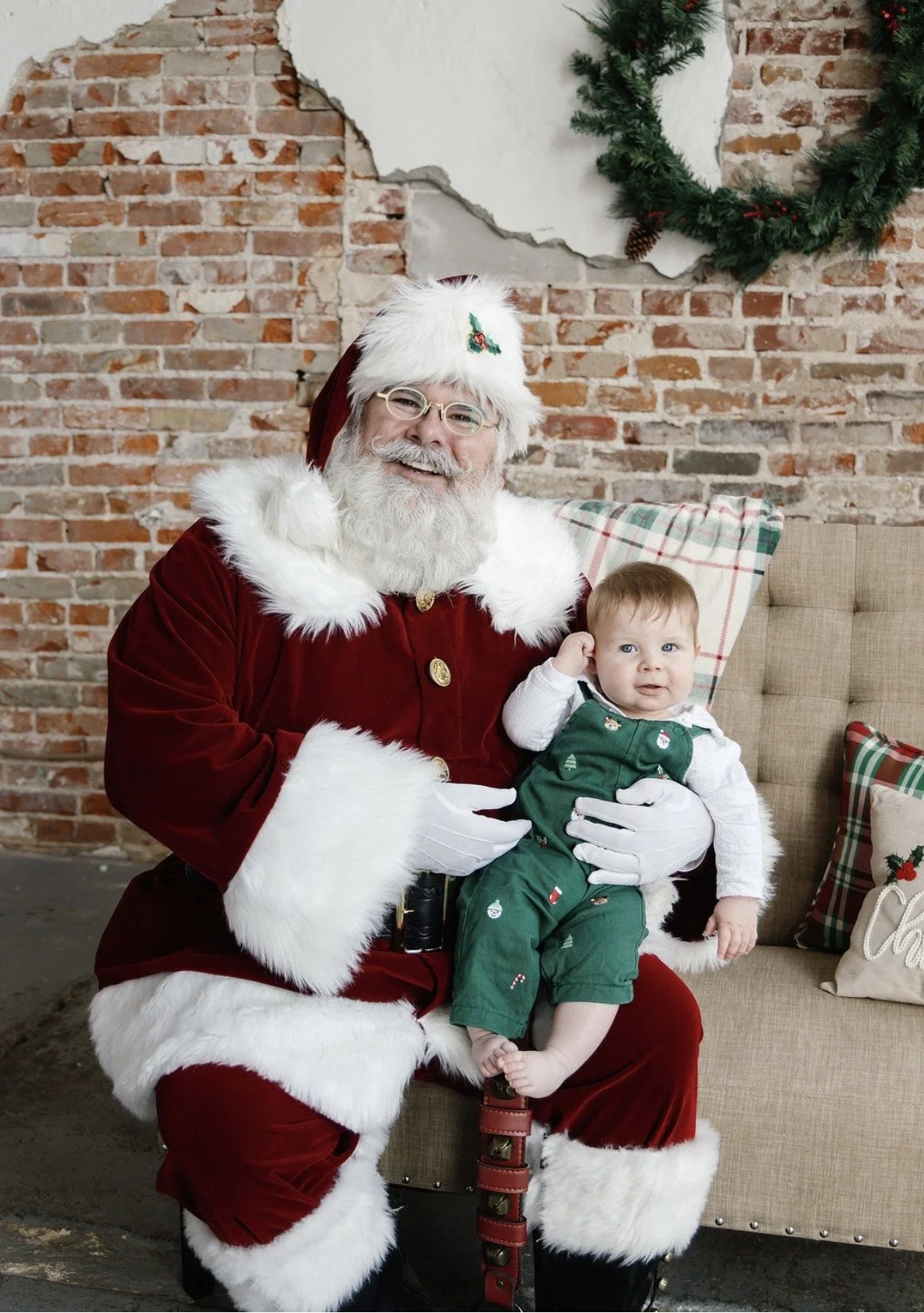 Santa Claus sitting on a chair holding a young child dressed in green holiday clothes, in front of a brick wall and Christmas wreath.