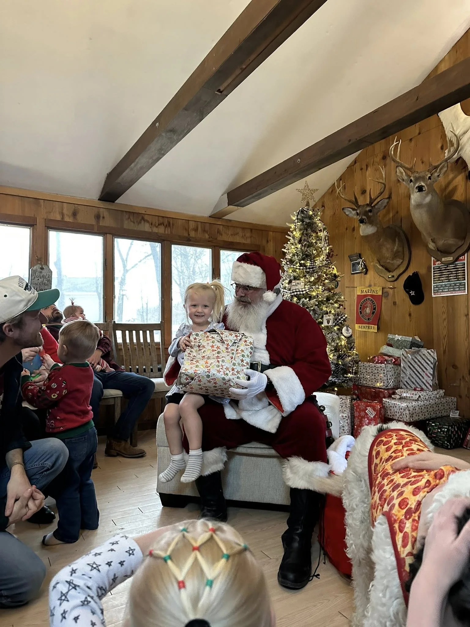 Children gathered around Santa Claus in a room decorated for Christmas, with a Christmas tree and wrapped presents in the background. Santa is holding a young girl on his lap, and there are other children and adults present. The room has wooden walls