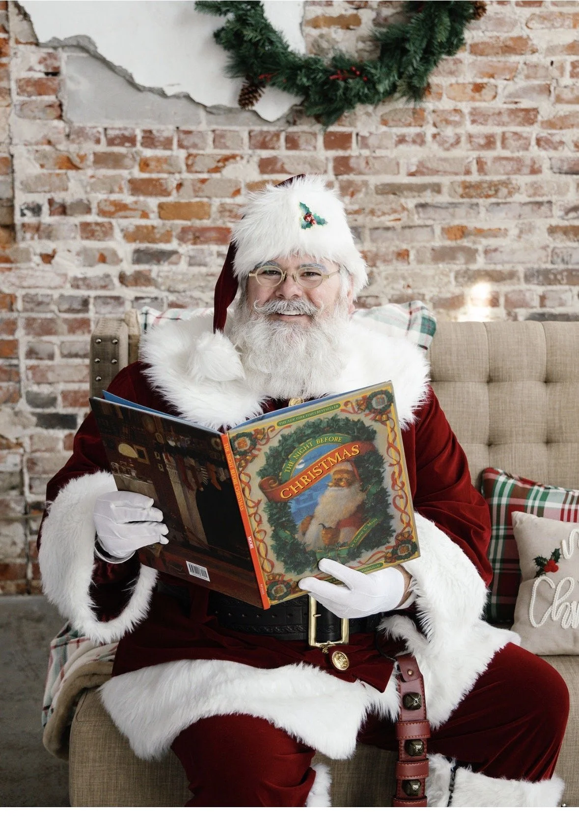 Santa Claus sitting on a beige chair, wearing a red velvet suit with white fur trim, reading a Christmas-themed book titled 'The Night Before Christmas', in front of a brick wall decorated with Christmas greenery and plaid pillows.