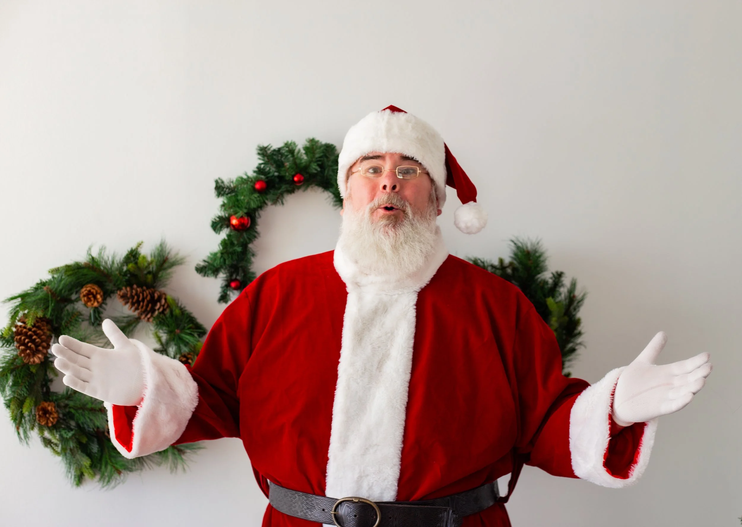 A man dressed as Santa Claus standing in front of a decorated Christmas backdrop with holiday greenery and red ornaments, gesturing with open hands.
