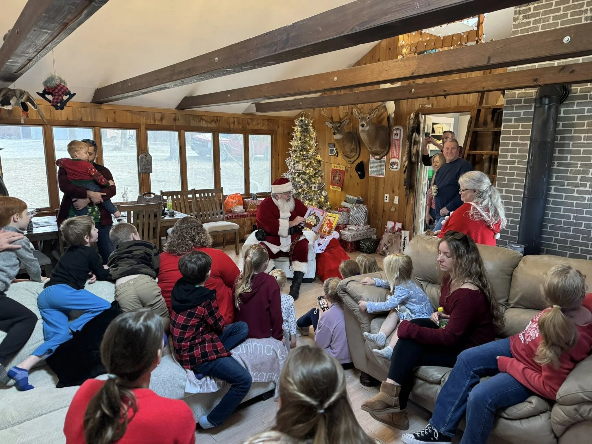 Multiple children and adults gathered in a cozy living room decorated with a small Christmas tree, holiday stockings, and festive decorations. Santa Claus is sitting on a couch reading a book to the children, who are sitting on the floor listening.