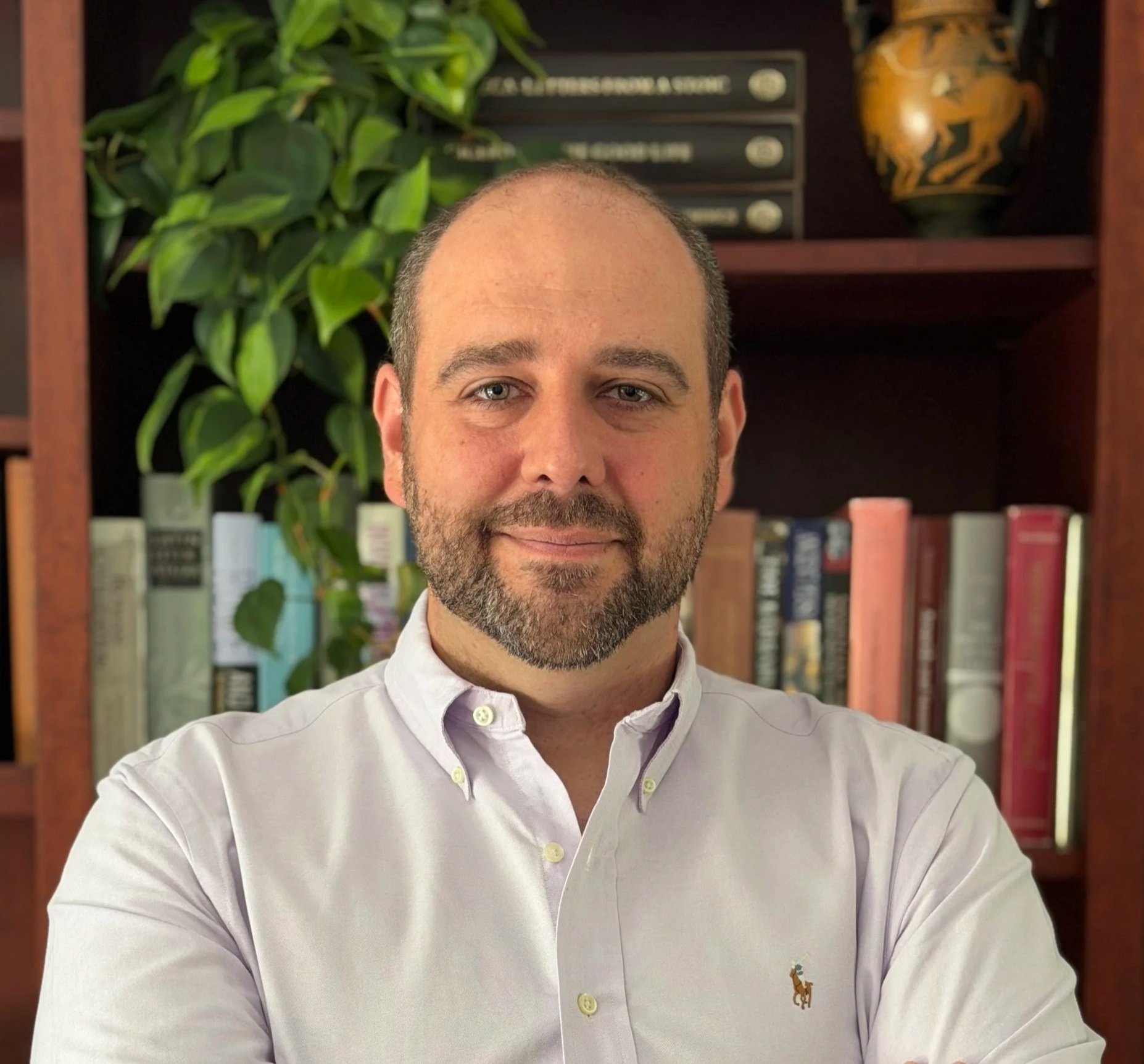Headshot of a bearded man with short brown hair, wearing a lavender button-down shirt, in front of a bookshelf with books, a plant, and decorative objects in the background.