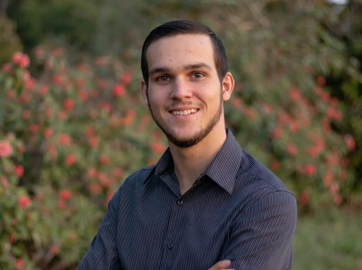 Portrait of a young man with dark hair and a beard, smiling, wearing a black striped shirt, outdoors with blurred pink flowers and green foliage in the background.