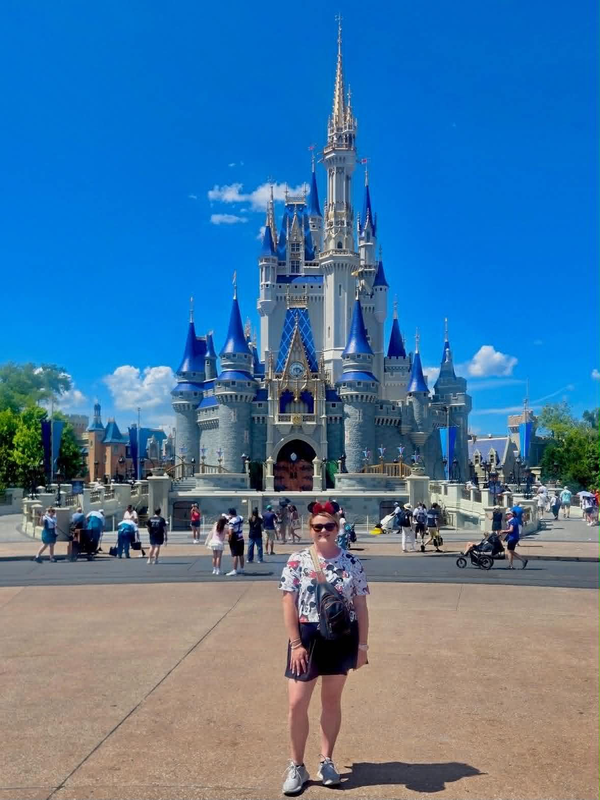 A woman standing in front of Cinderella's Castle at a Disney theme park on a bright sunny day, with people walking around and a clear blue sky in the background.