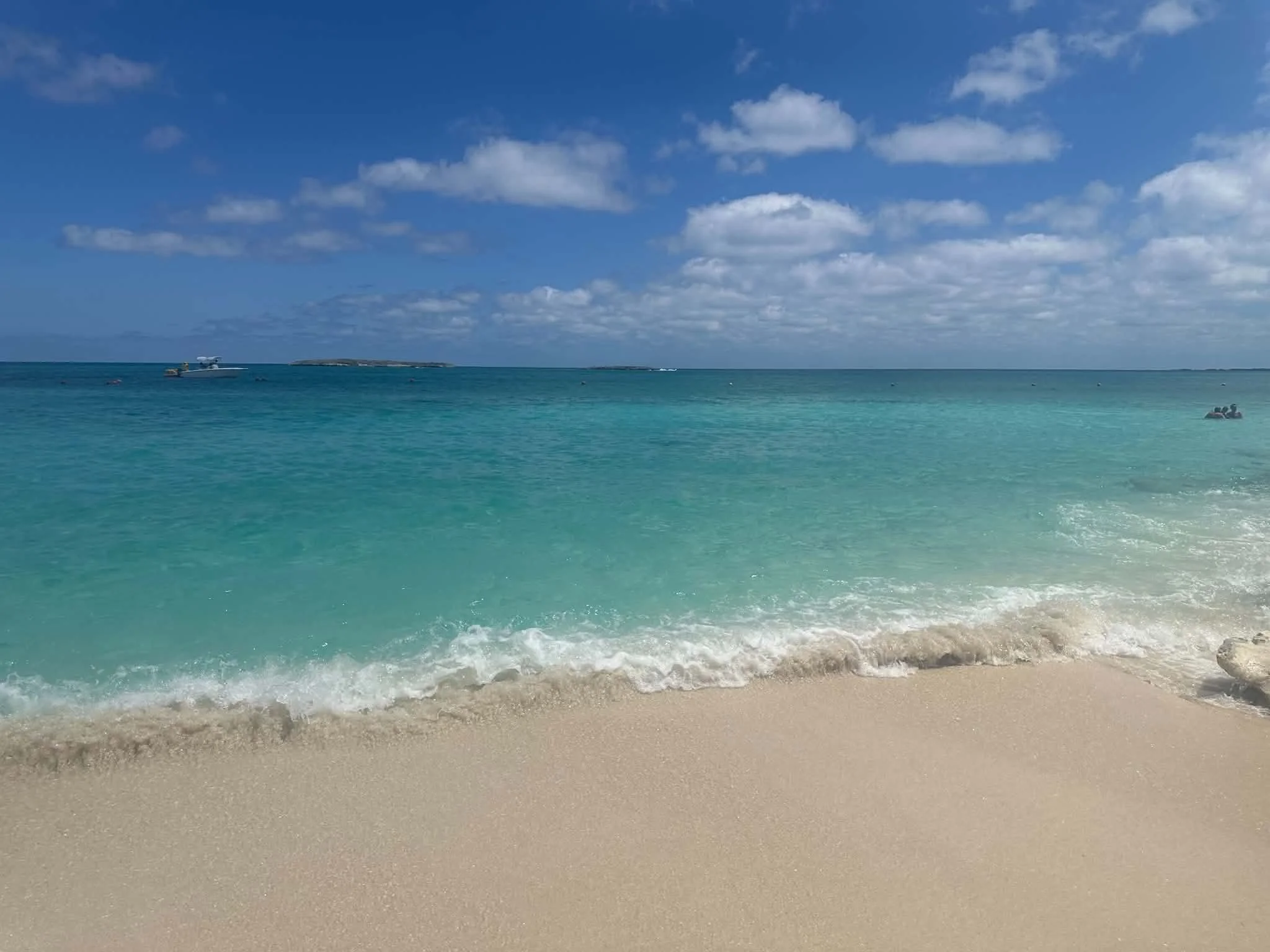 A sandy beach with turquoise water, a boat in the distance, and a partly cloudy sky.