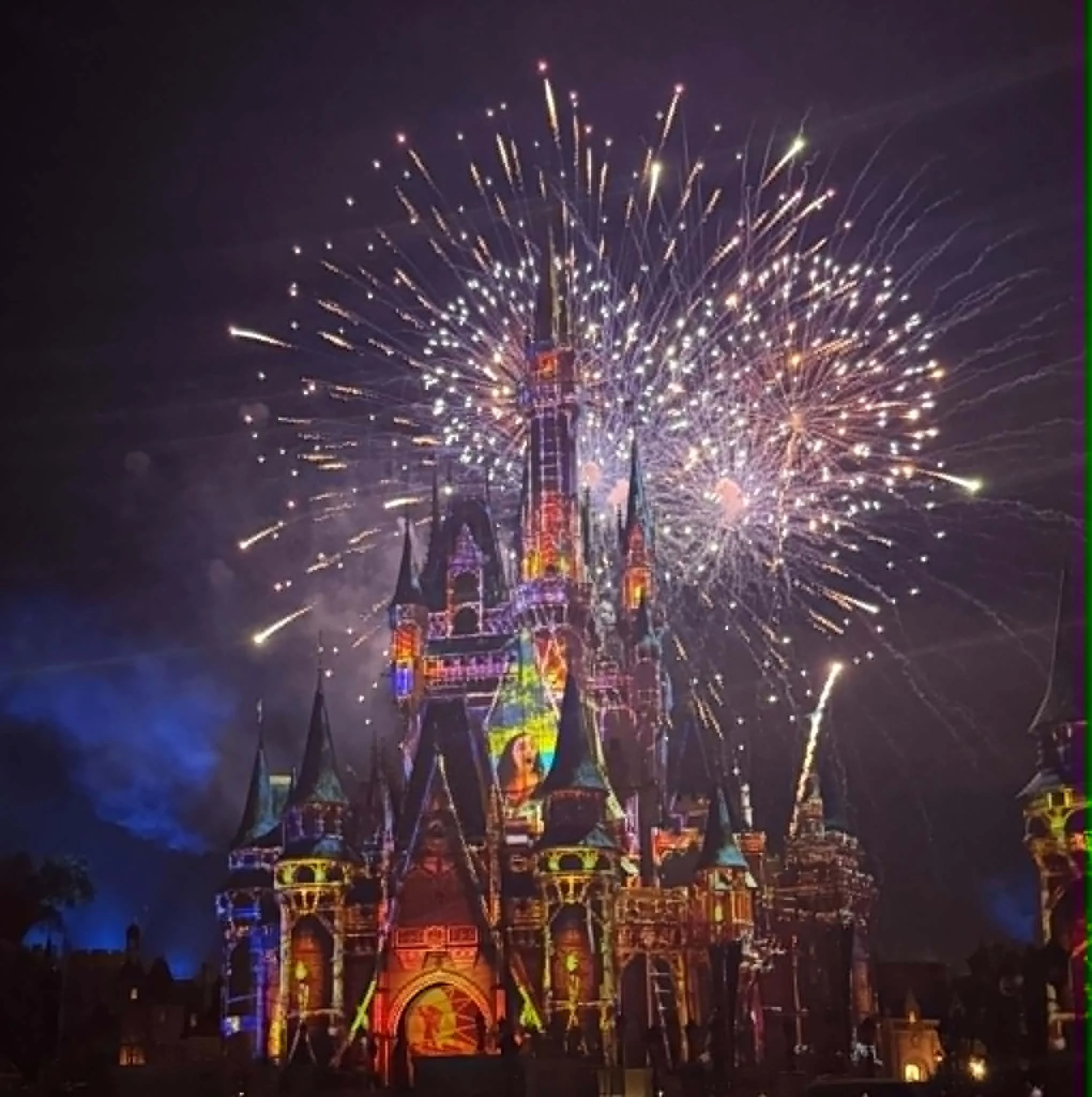Nighttime fireworks display above a brightly lit amusement park attraction, with colorful lights and intricate metal framework.