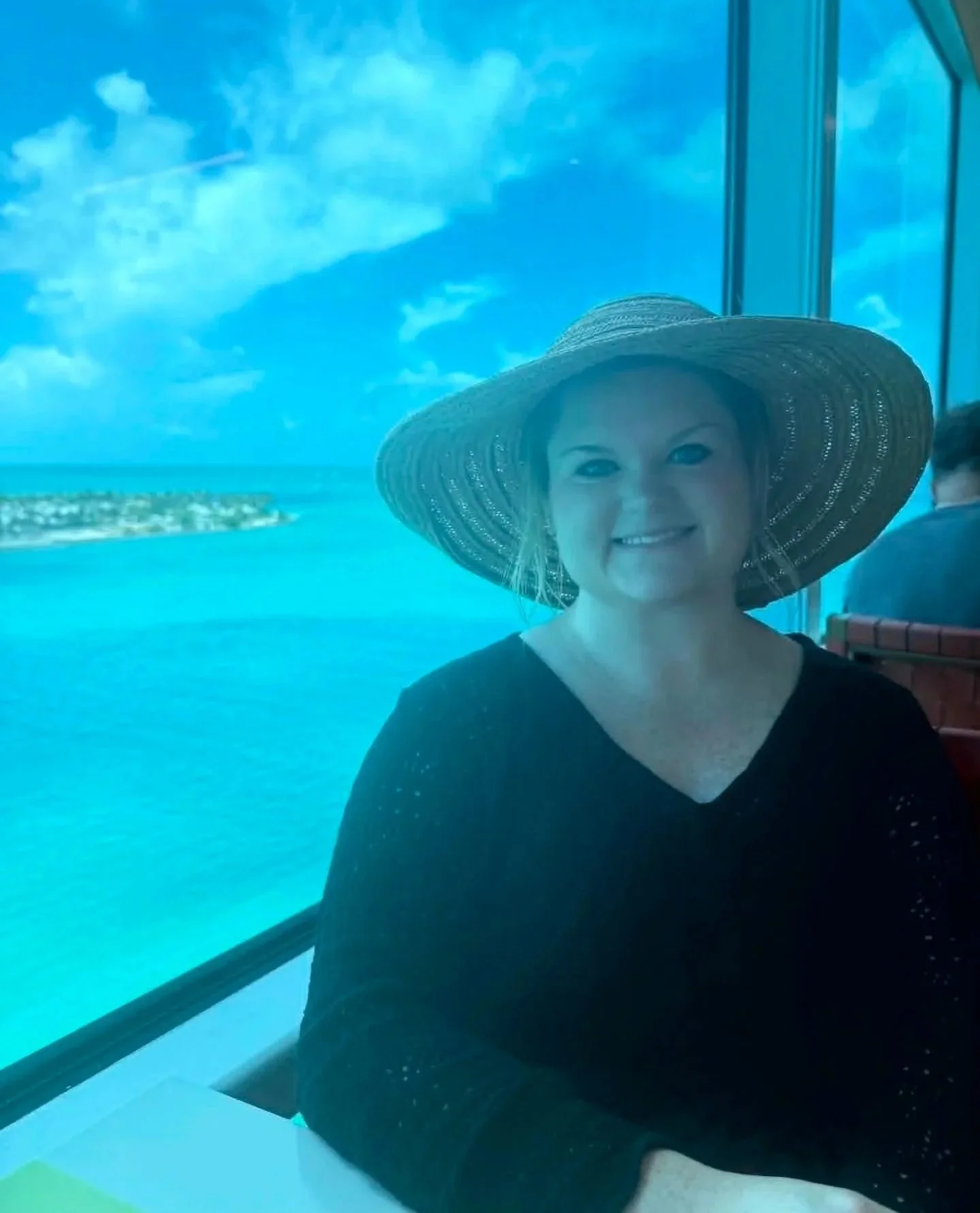 A woman smiling while wearing a large straw sunhat, sitting by a window overlooking an ocean and a small island, with a blue sky and clouds.
