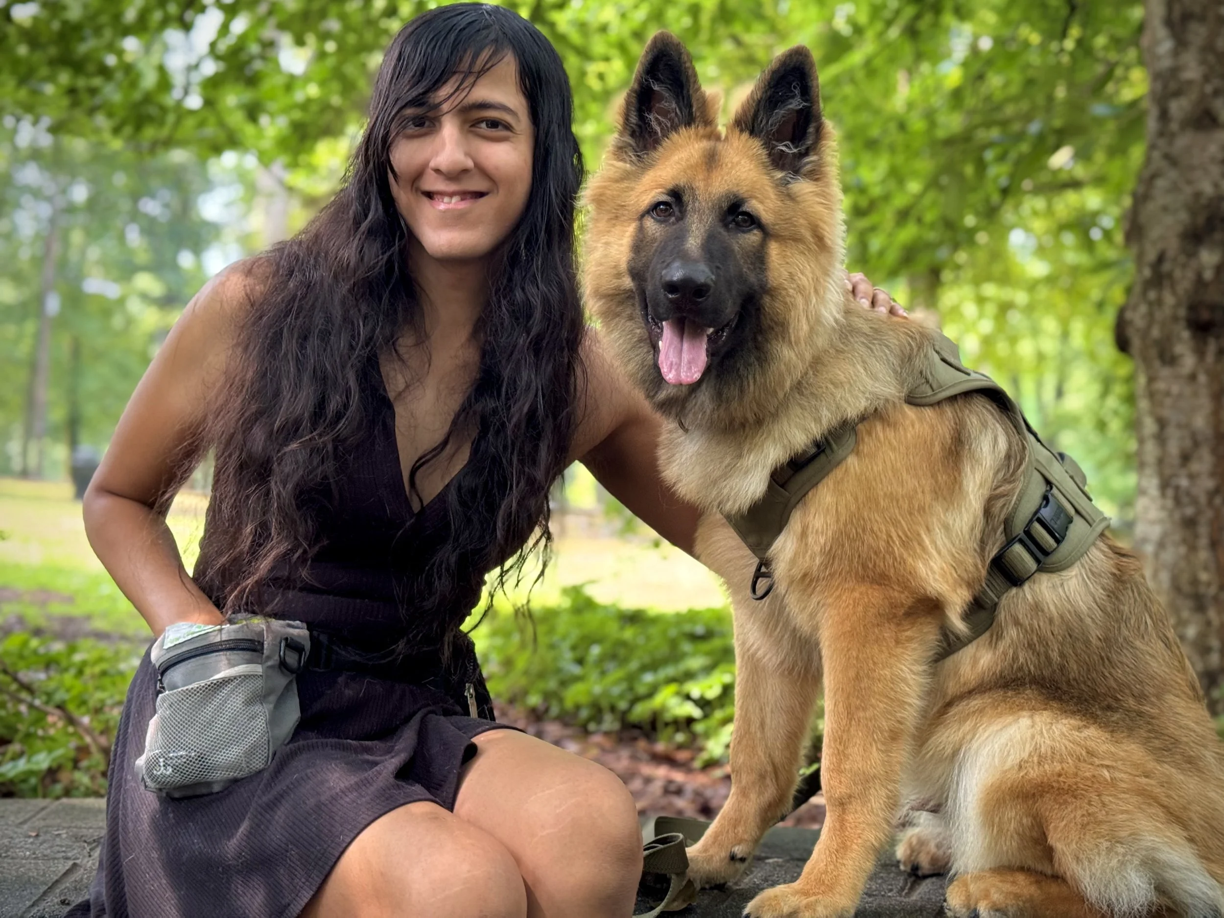 A person with long dark hair and a dog with a tan coat and black face markings, both in a wooded outdoor area.