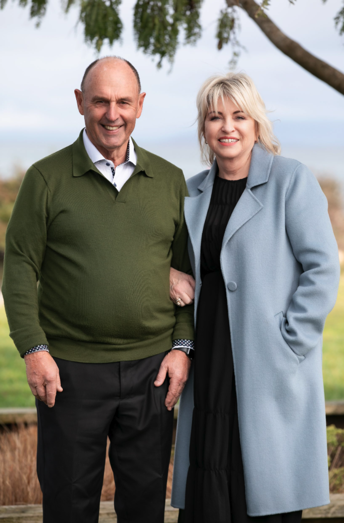 A smiling middle-aged man and woman standing outdoors under a tree on a cloudy day, holding hands.
