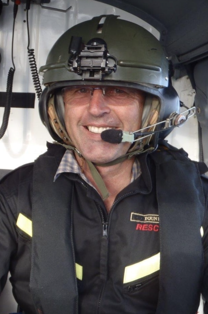 John Funnell wearing a rescue  helmet with attached communication gear, headset, and a rescue jacket inside an aircraft or helicopter.