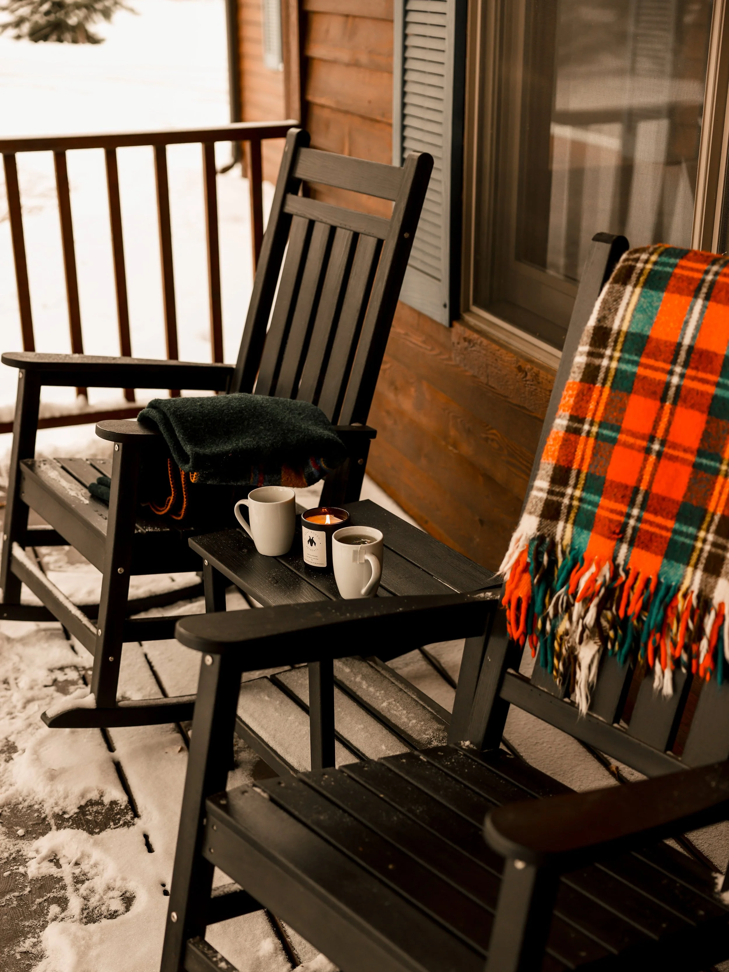 Front porch with two black wooden rocking chairs, one with a folded plaid blanket and the other with a black hat and zipped jacket, on a snow-covered wooden deck in winter.