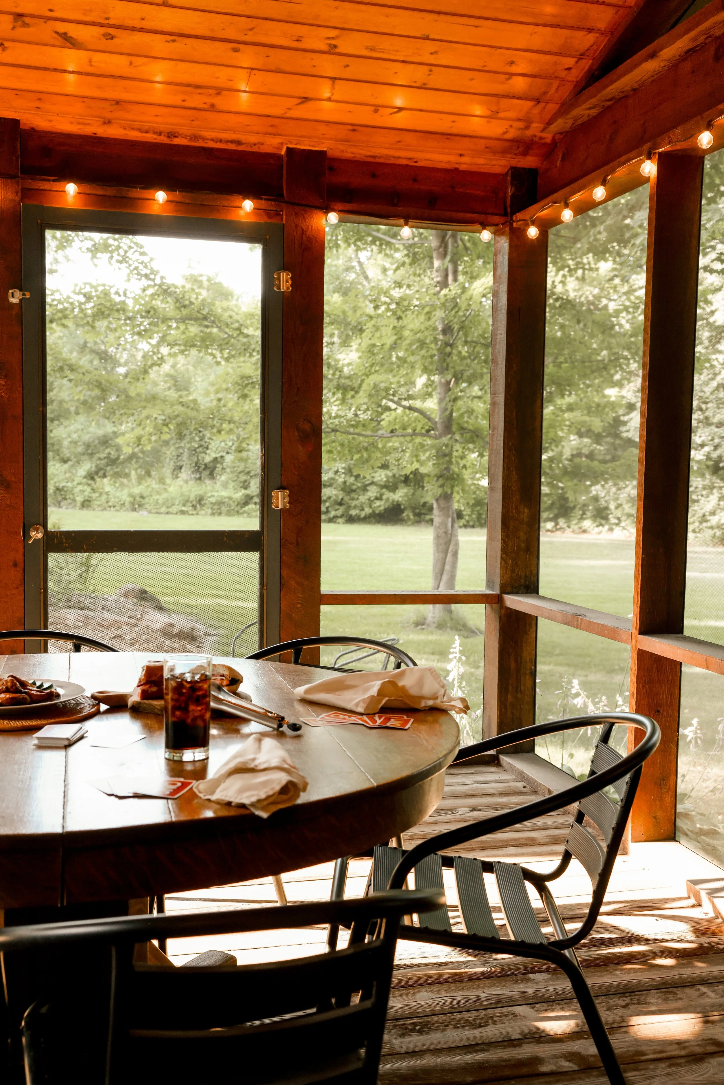 A wooden screened porch with a table set for a meal, featuring plates of food and a glass of dark soda, overlooking lush green trees outside. String lights hang from the ceiling.