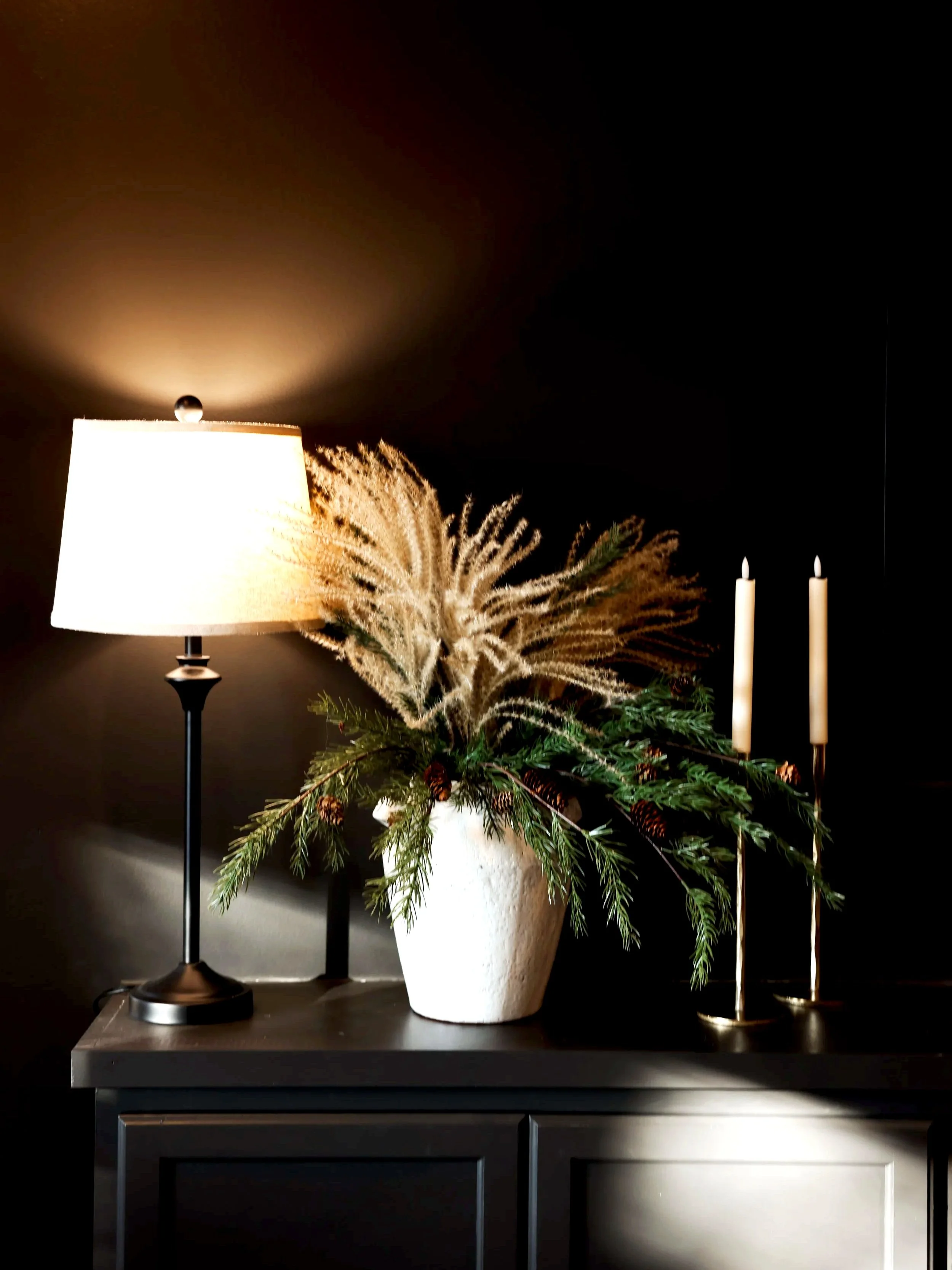 A decorated holiday table with a white vase holding pine branches and pinecones, a lamp with a beige shade casting a warm glow, and two tall white candles on gold holders against a dark background.